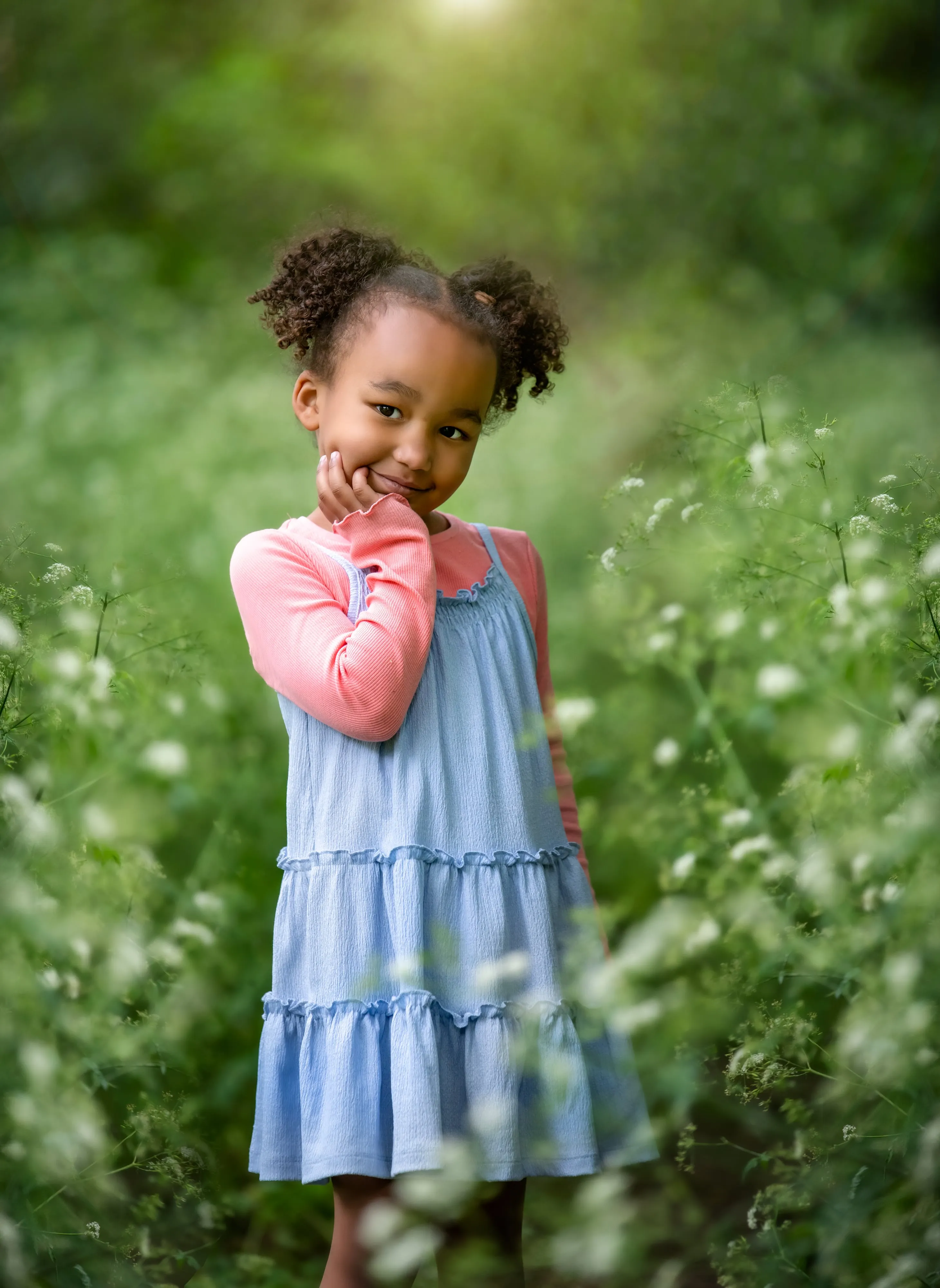 Young girl with curly hair in pale-blue tiered dress smiles shyly among Queen Anne's lace wildflowers in soft summer light