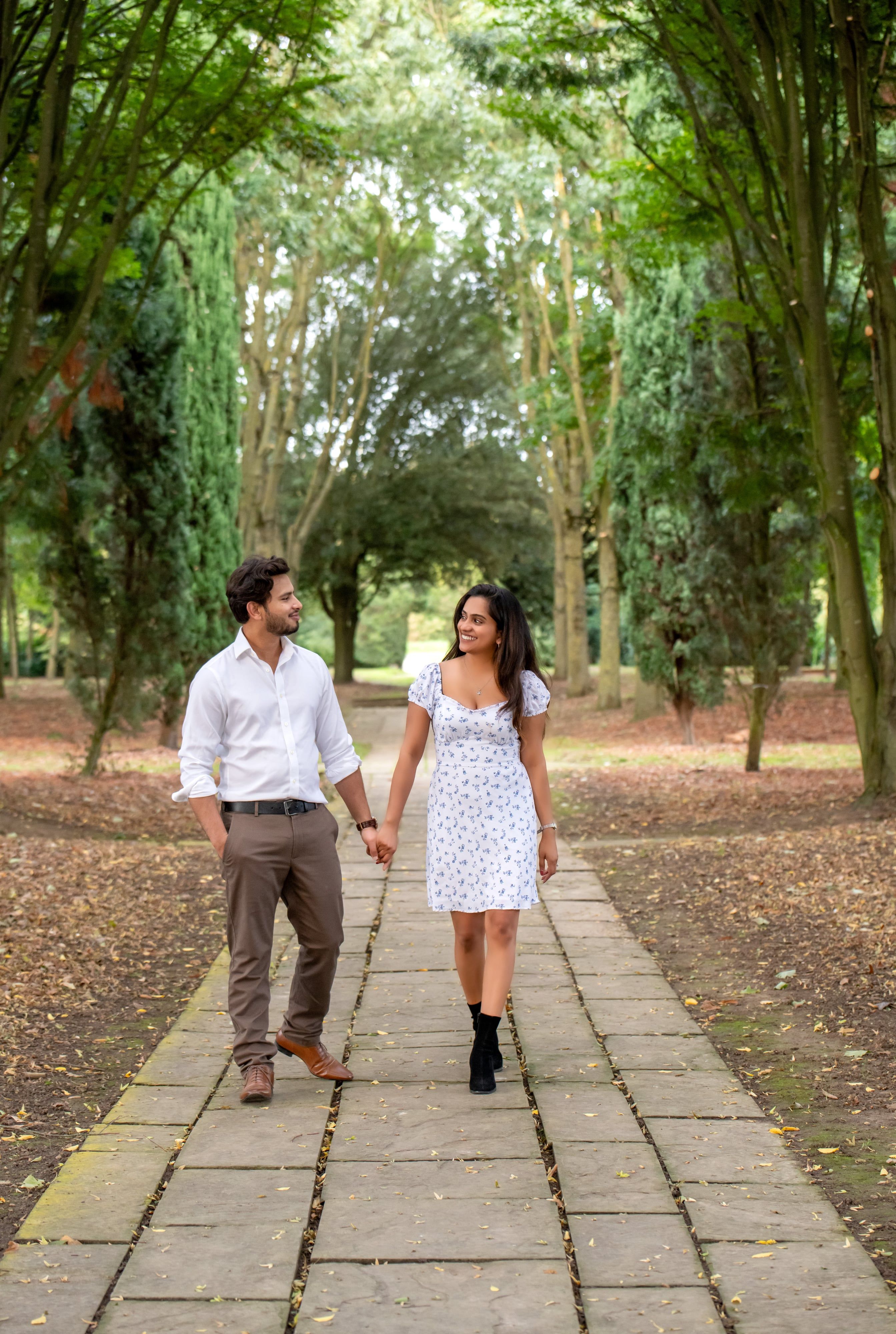 Couple in white shirt and floral dress walk hand-in-hand along a tree-lined parkland path