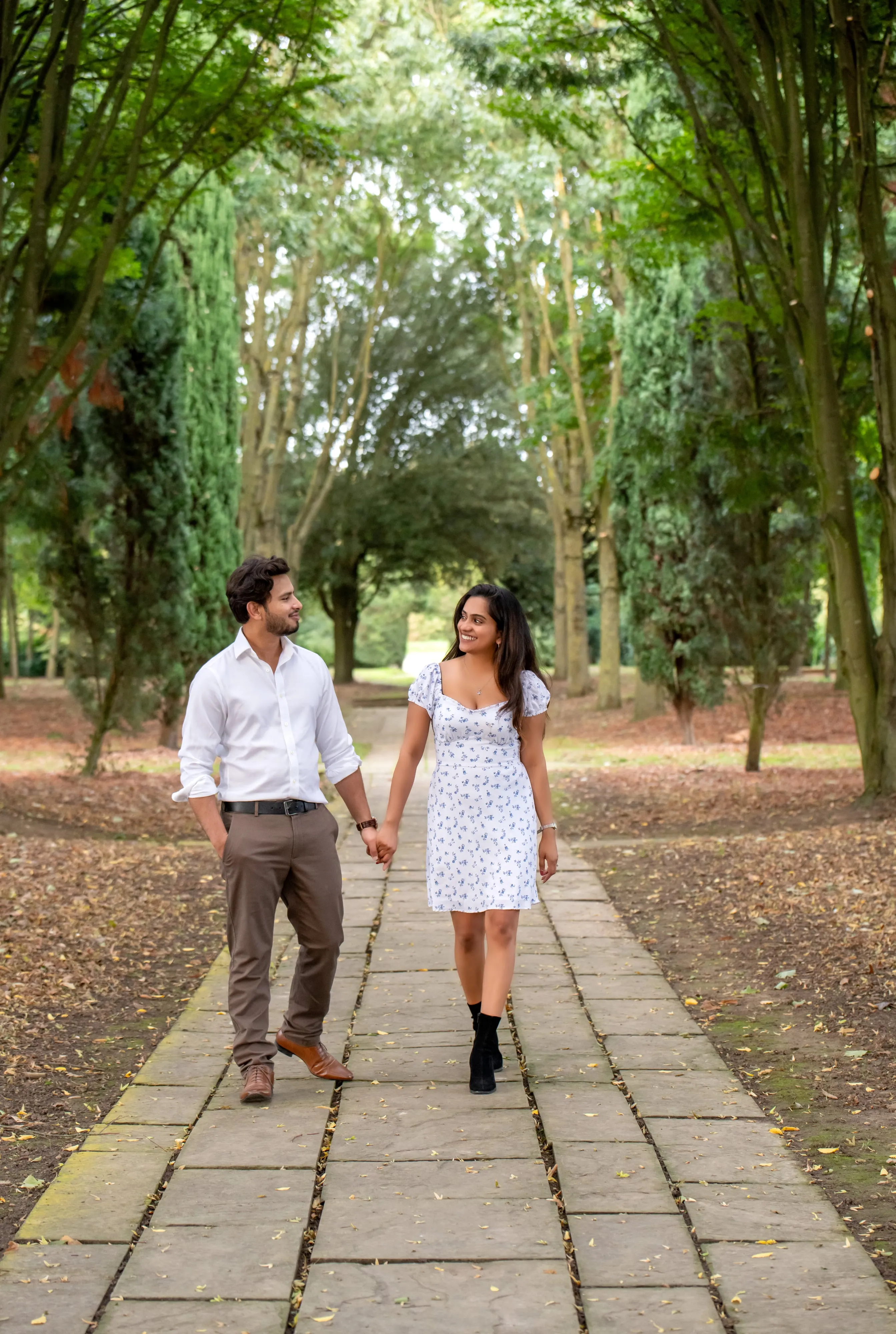 Couple in white shirt and floral dress walk hand-in-hand along a tree-lined parkland path