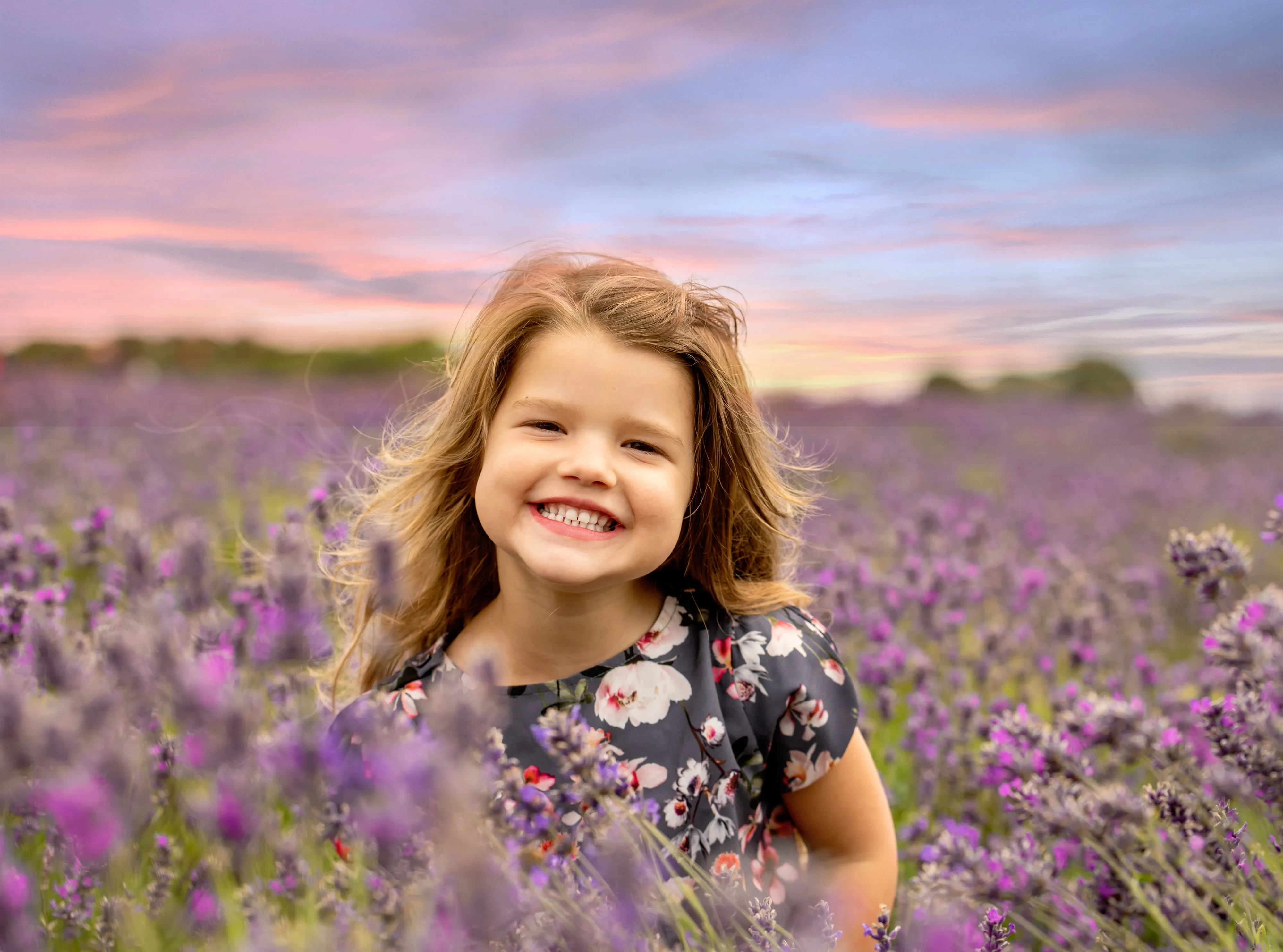Young girl in floral dress beams in a lavender field at sunset, pink and lilac sky behind her
