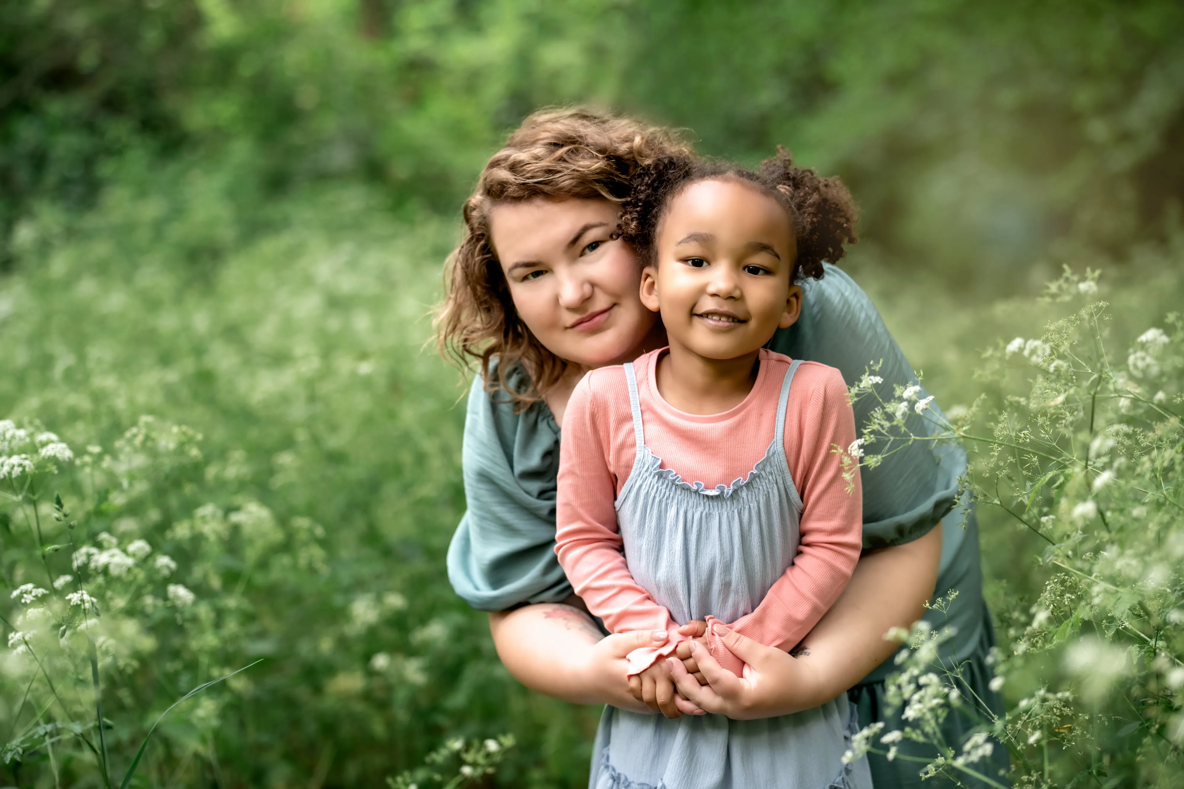 Mother in sage wrap dress hugs young daughter in pink and grey pinafore amid summer cow parsley, soft golden light