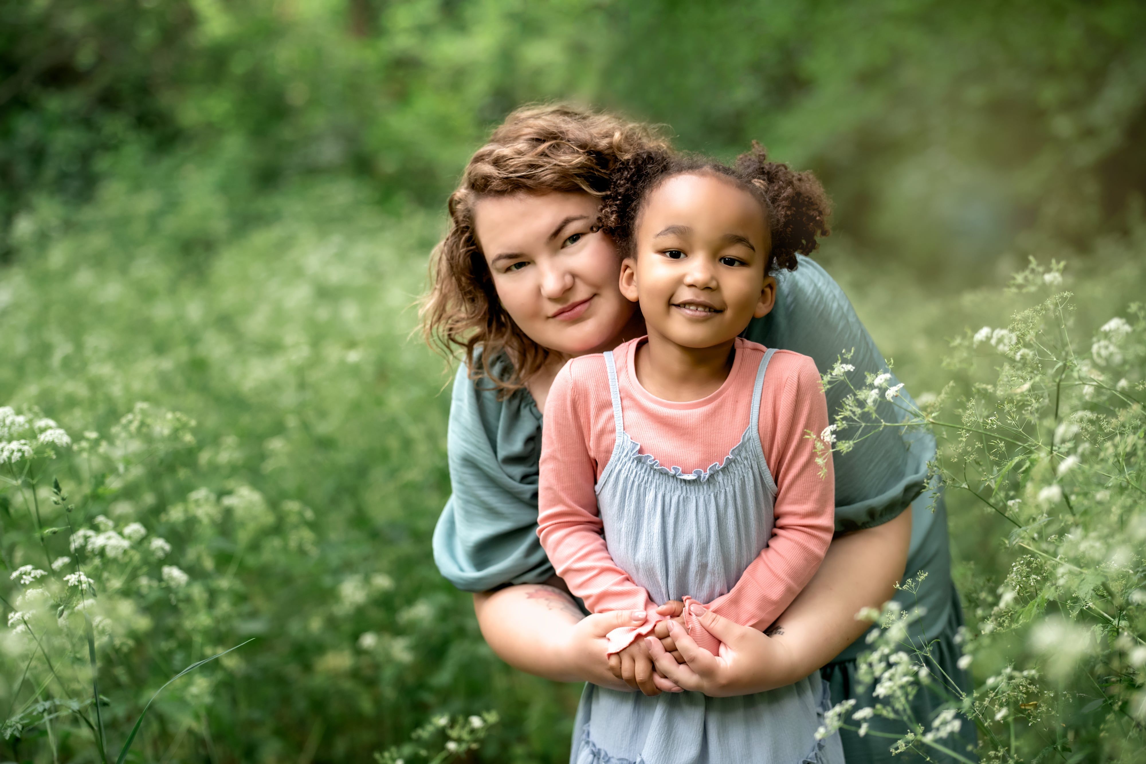 Mother in sage wrap dress hugs young daughter in pink and grey pinafore amid summer cow parsley, soft golden light