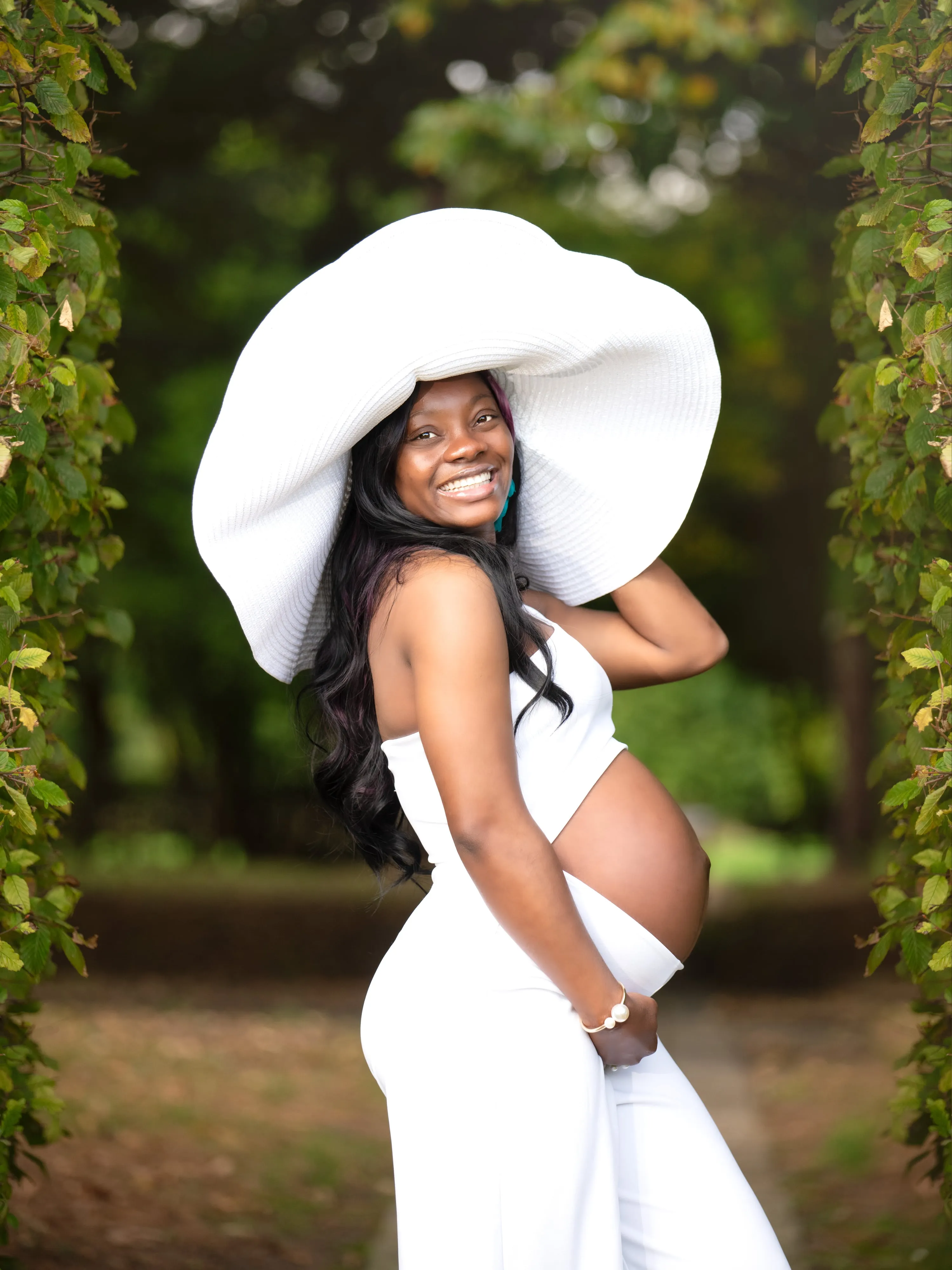 Maternity portrait — expecting mum in cropped white outfit and oversized sun hat smiles in a lush green archway