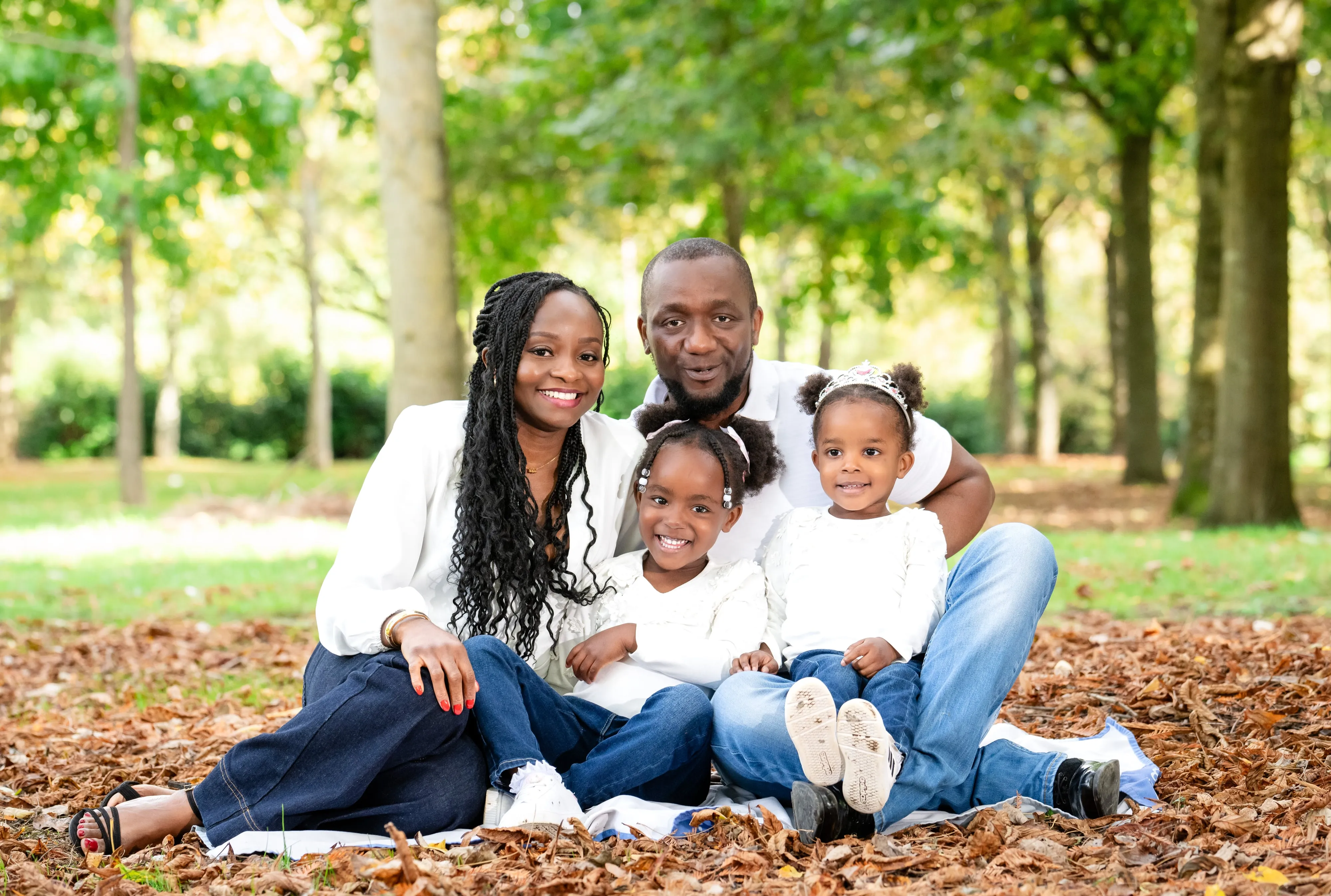 Family of four sit on a blanket in autumn leaves under mature trees — parents and two little girls in soft cream and denim
