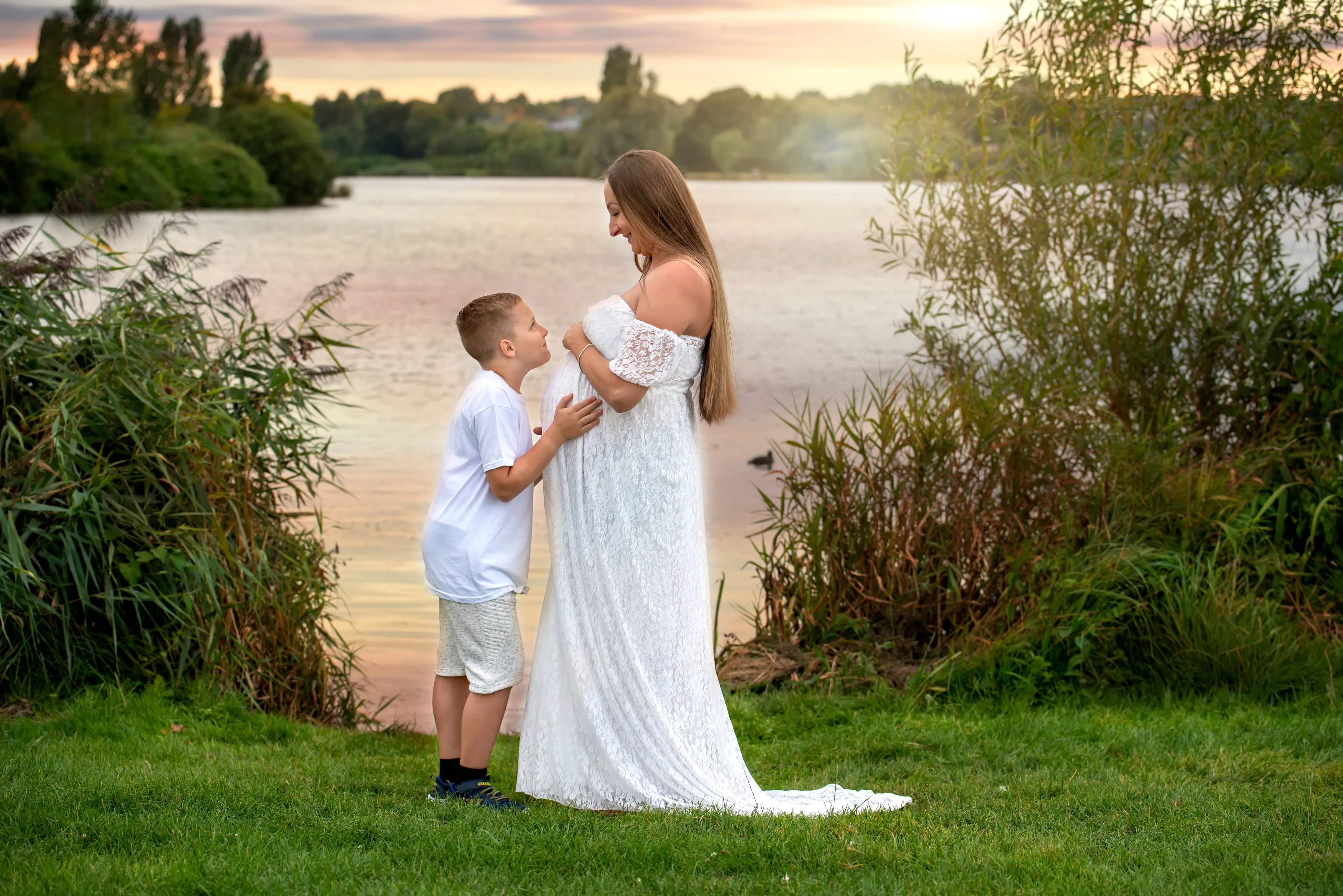 Maternity session at sunset — young son leans to kiss the bump as his mum smiles in a flowing white gown by the lake