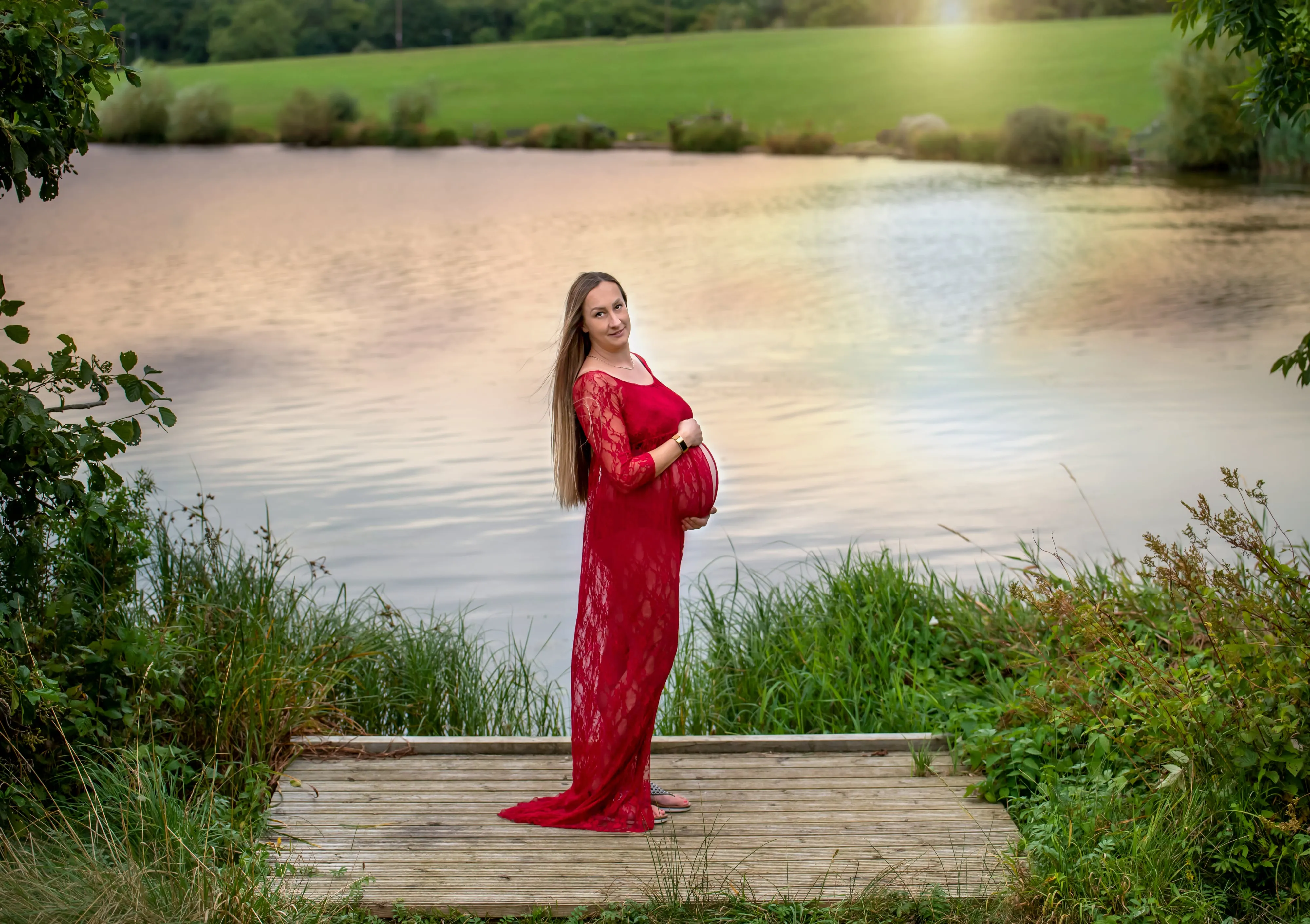 Maternity portrait at lakeside — expecting mum in flowing red lace gown cradles her bump on a wooden walkway at golden hour