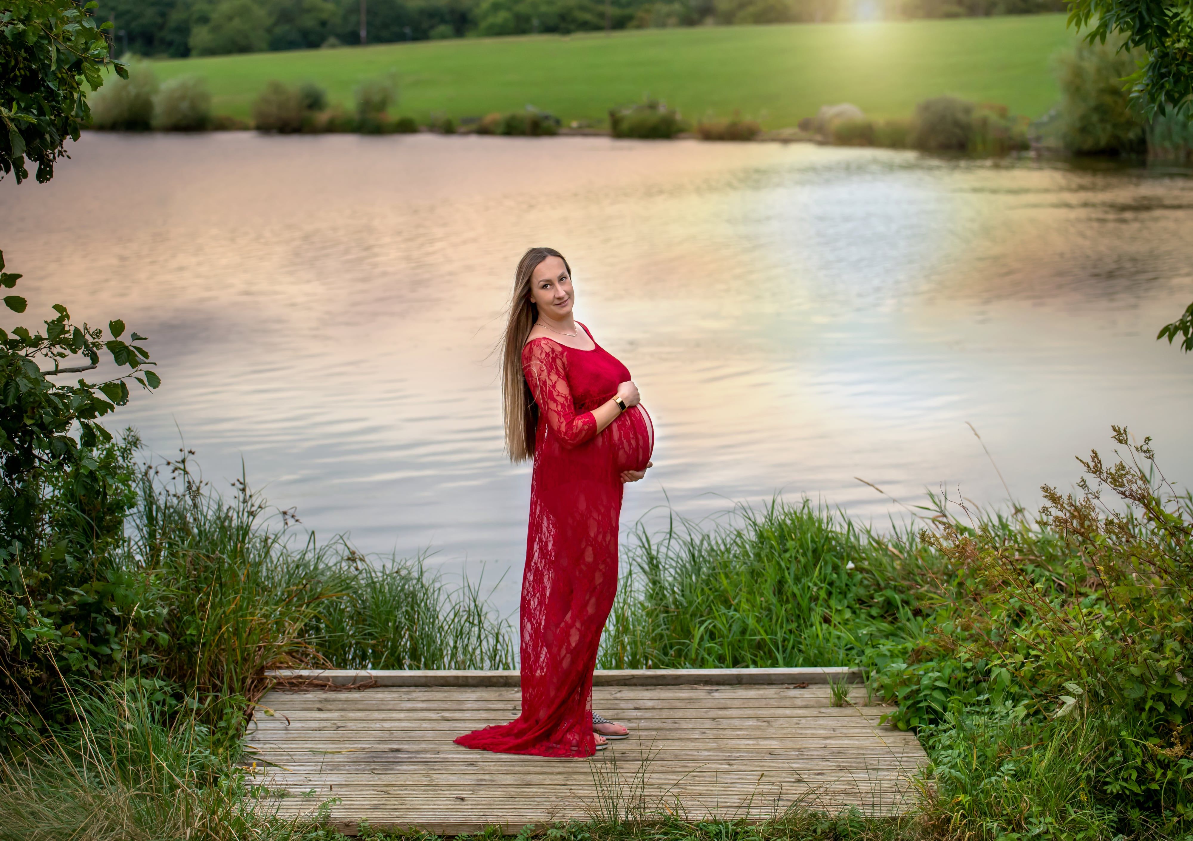 Maternity portrait at lakeside — expecting mum in flowing red lace gown cradles her bump on a wooden walkway at golden hour