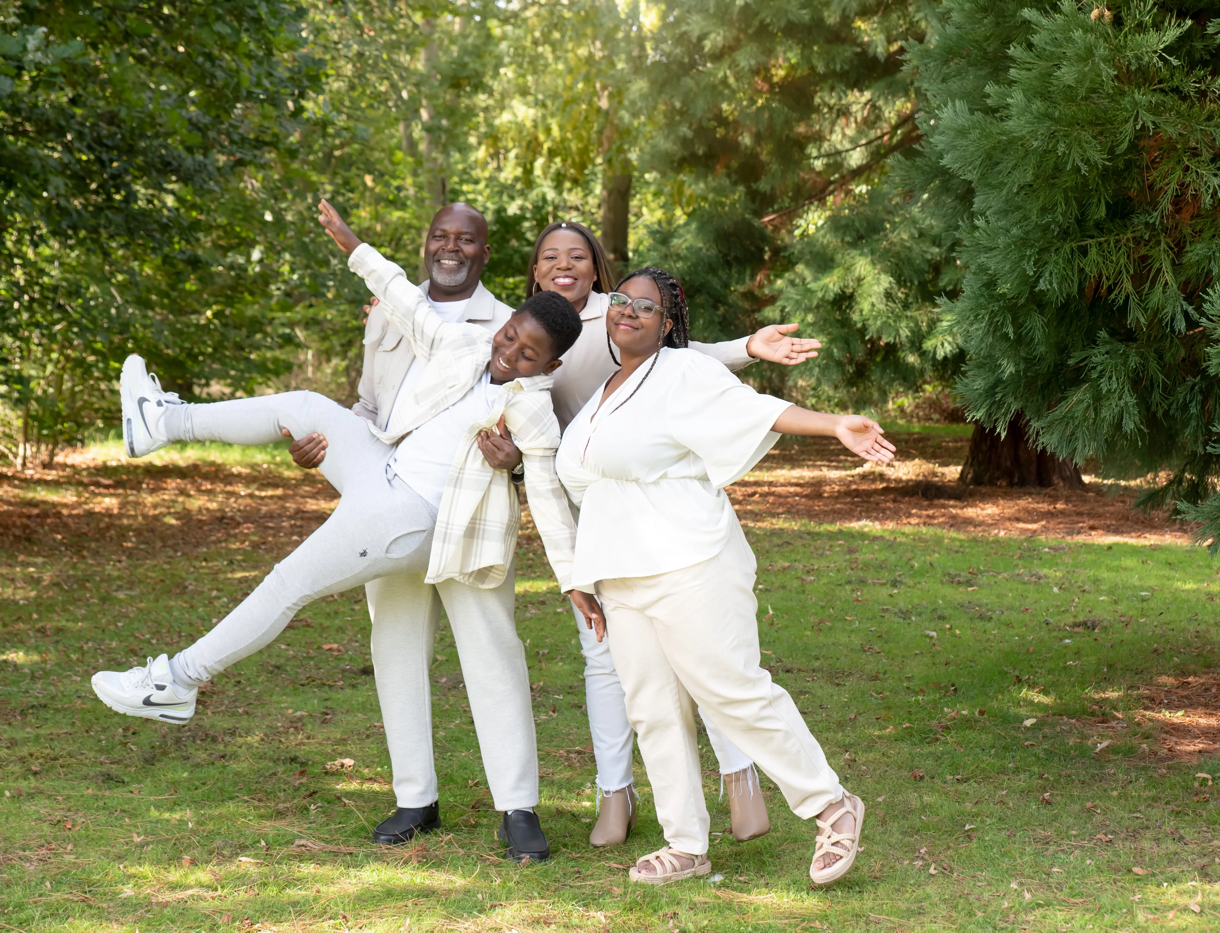 Family of three burst into a playful jump in matching cream outfits on a sunlit parkland lawn