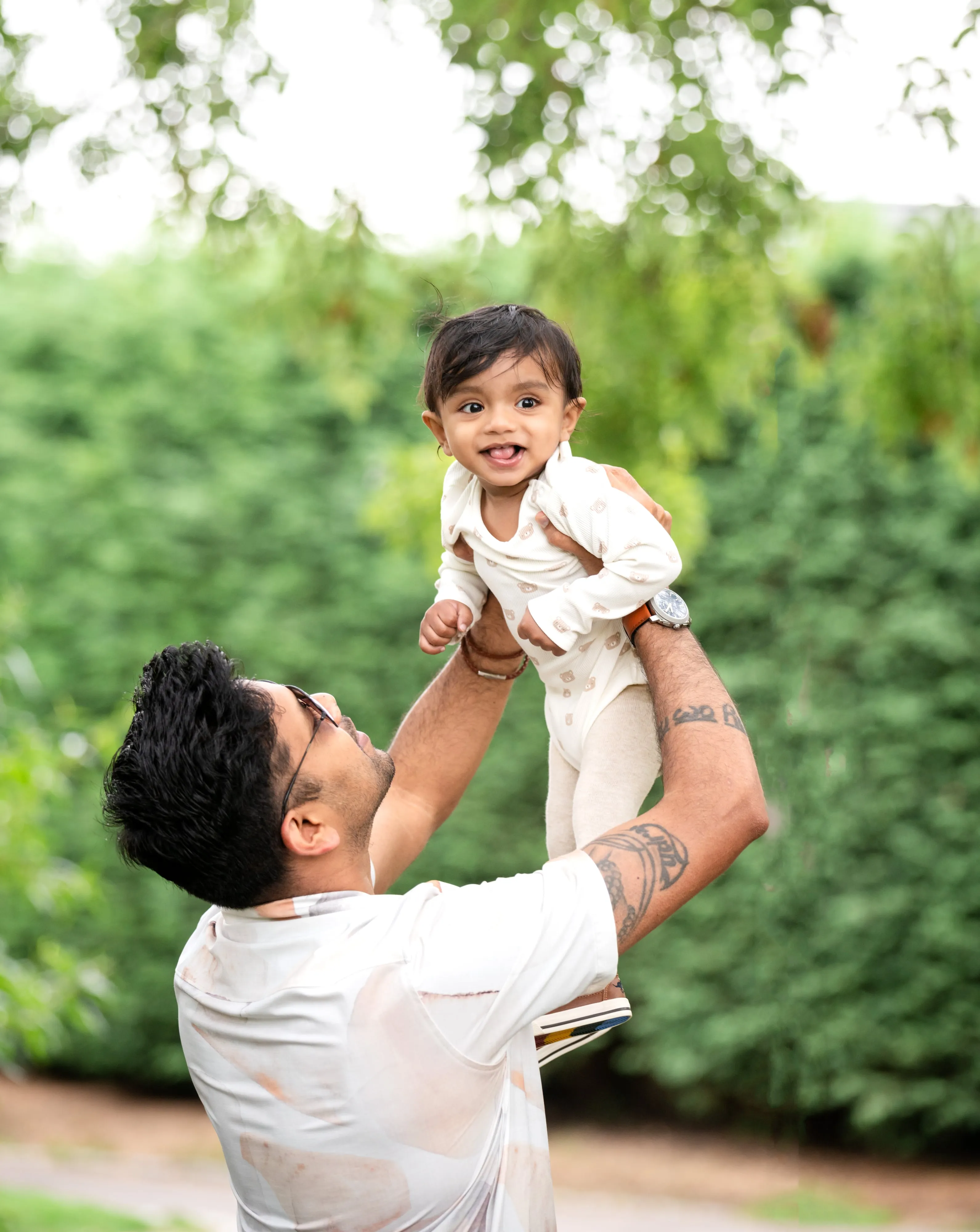 Father lifts laughing baby girl in cream romper above his head against a green hedge, late summer light