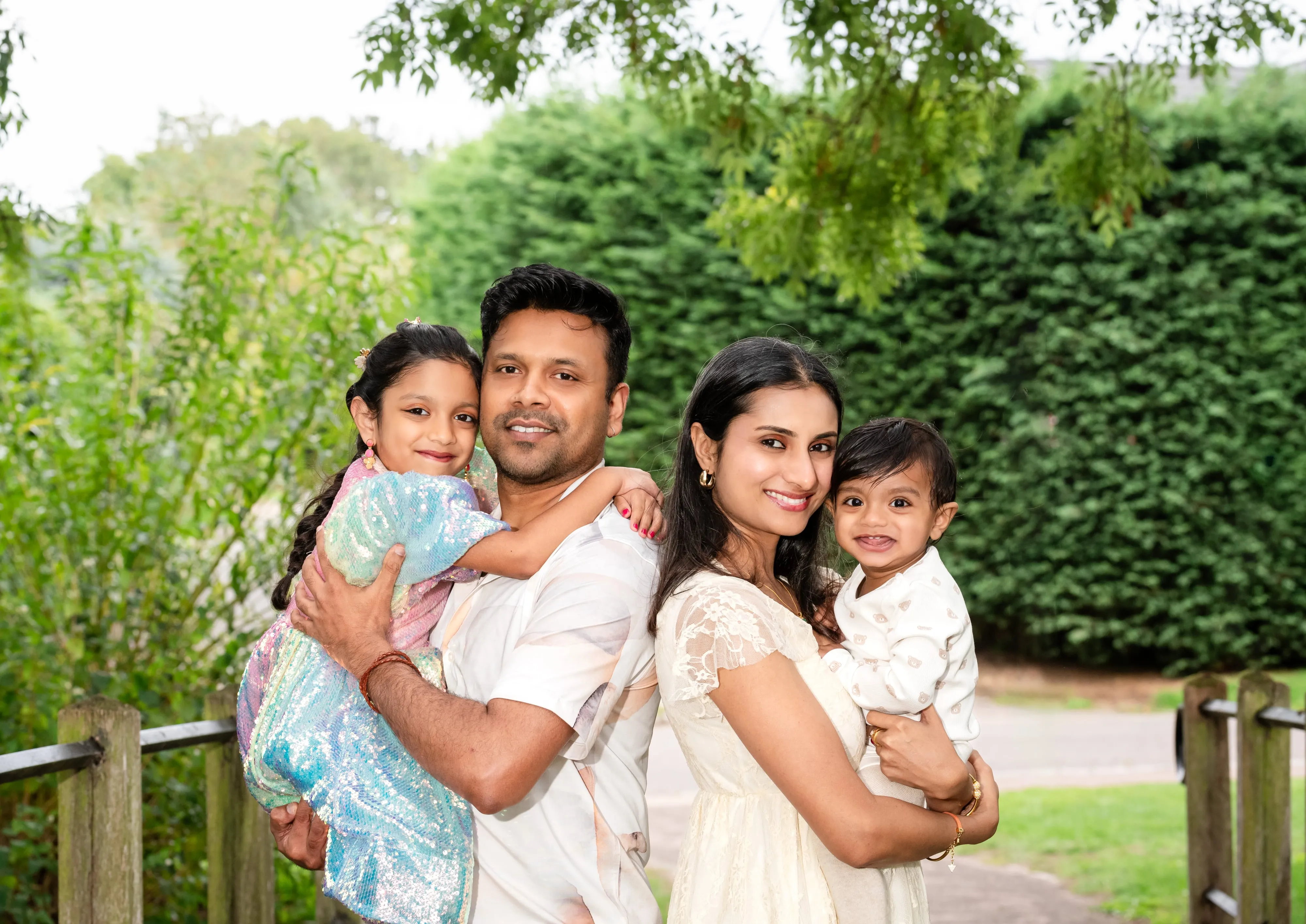 Family of four pose on a wooden footbridge against a clipped hedge — parents in white, two daughters in pastel dresses