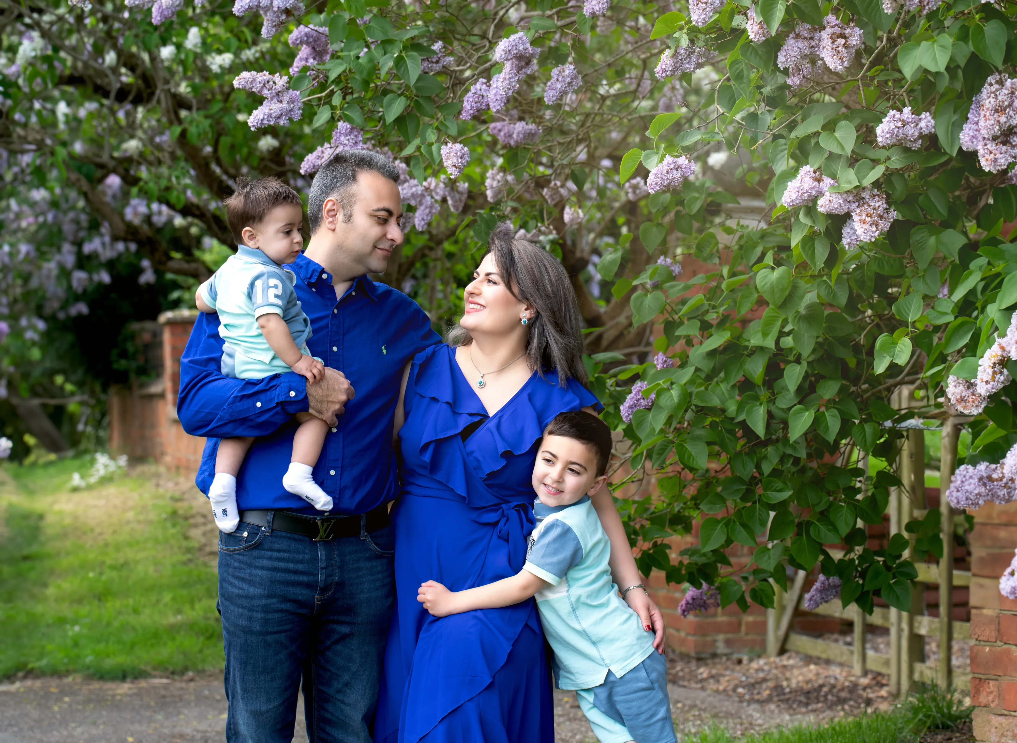 Family of four with two young boys laugh together in front of a lilac shrub in full bloom