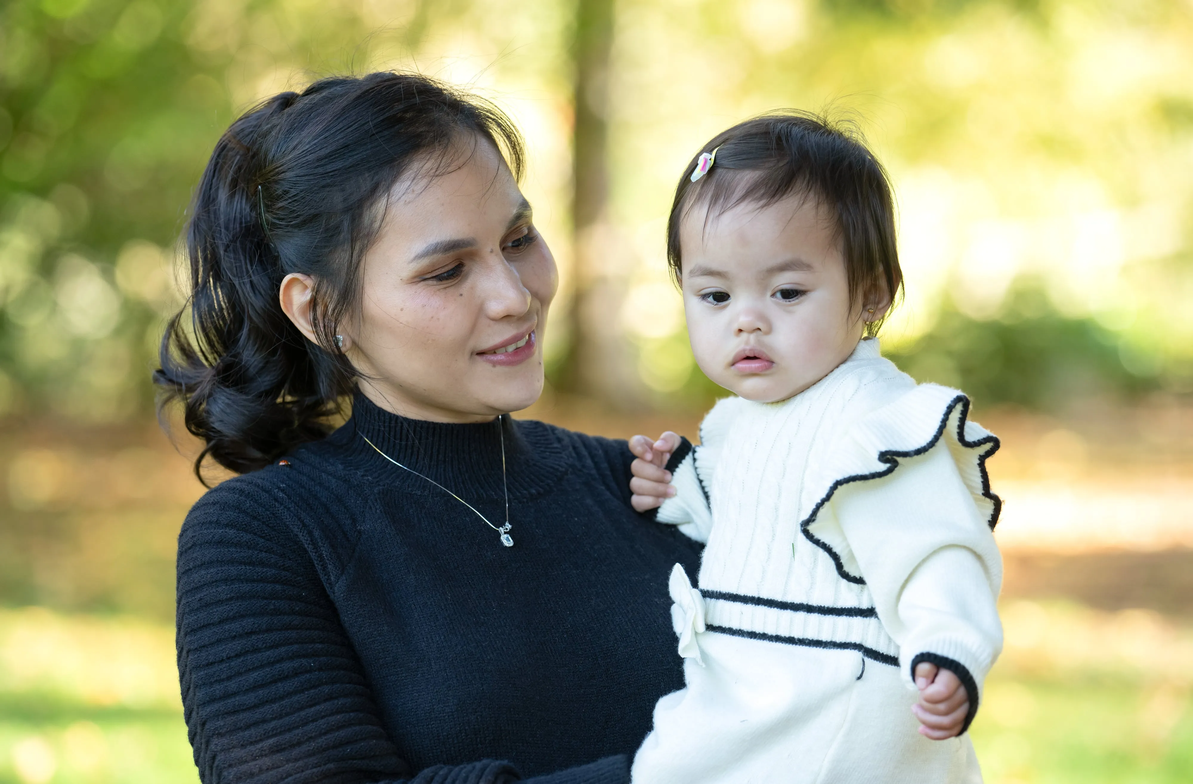 Mother in black knit holds her baby daughter in cream ruffled jumper in dappled autumn parkland light