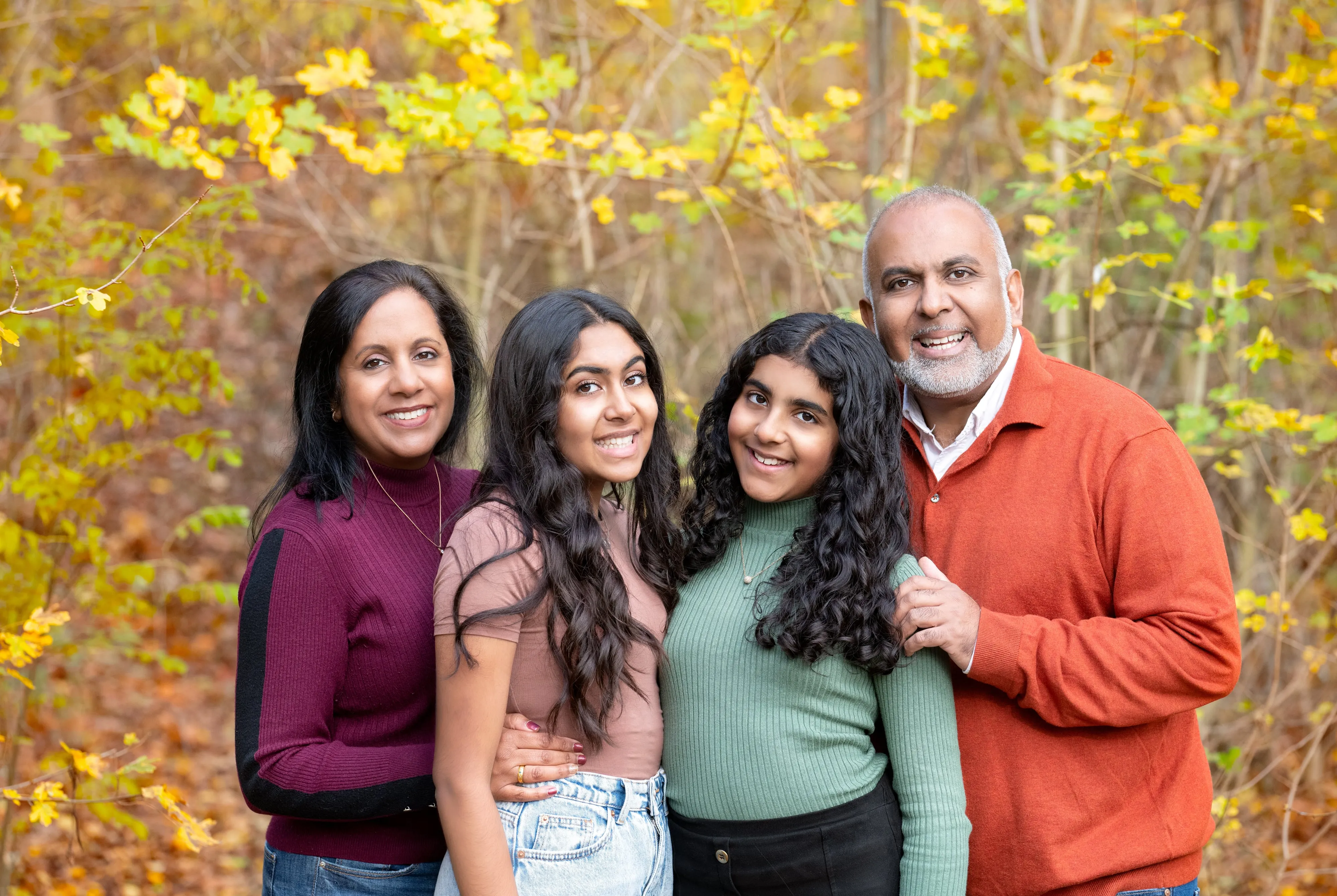 Family of four with two teenage daughters in earth-tone knits gather close among golden-yellow autumn maples