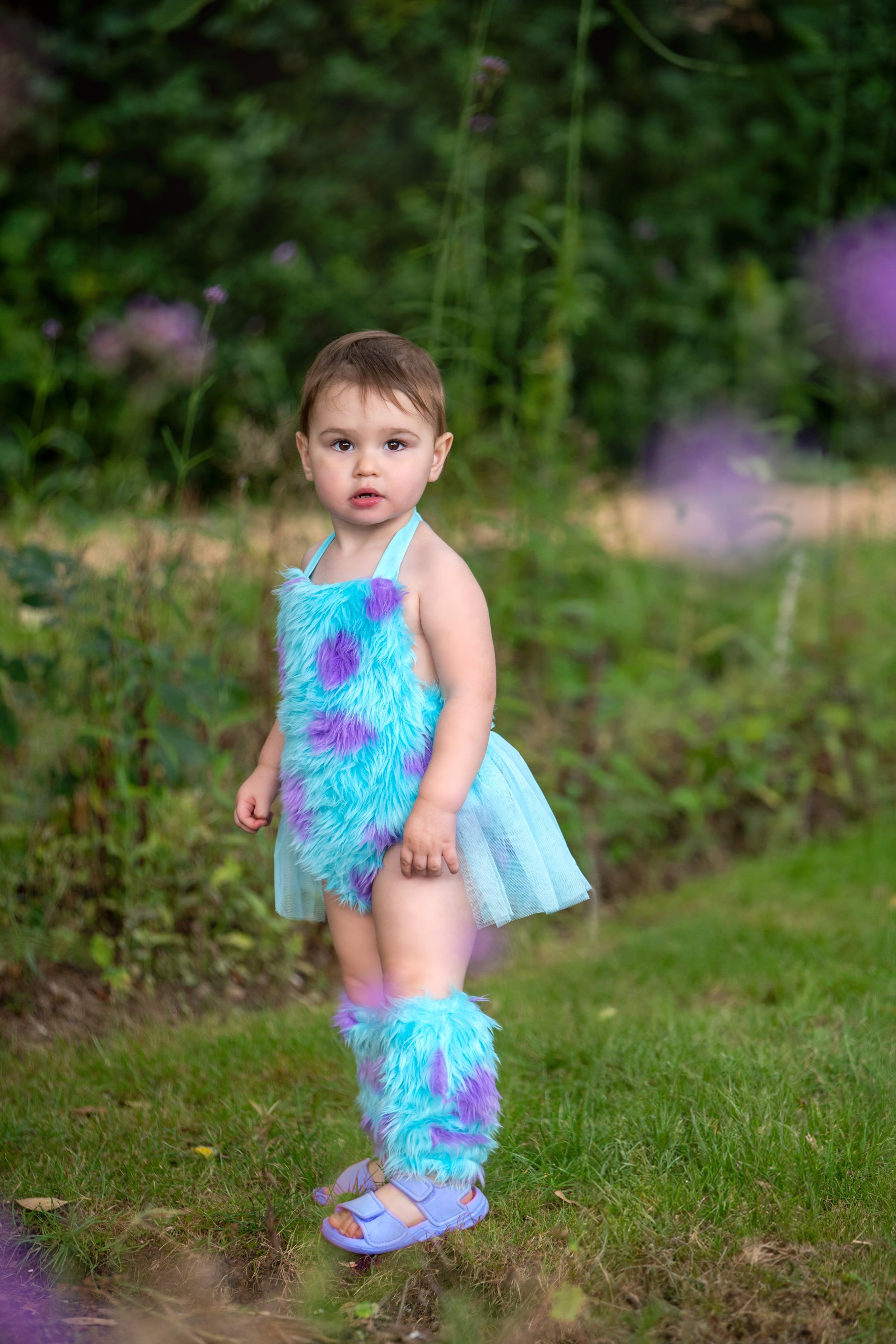 Toddler poses confidently in a fluffy turquoise-and-purple character outfit in a sunlit summer meadow