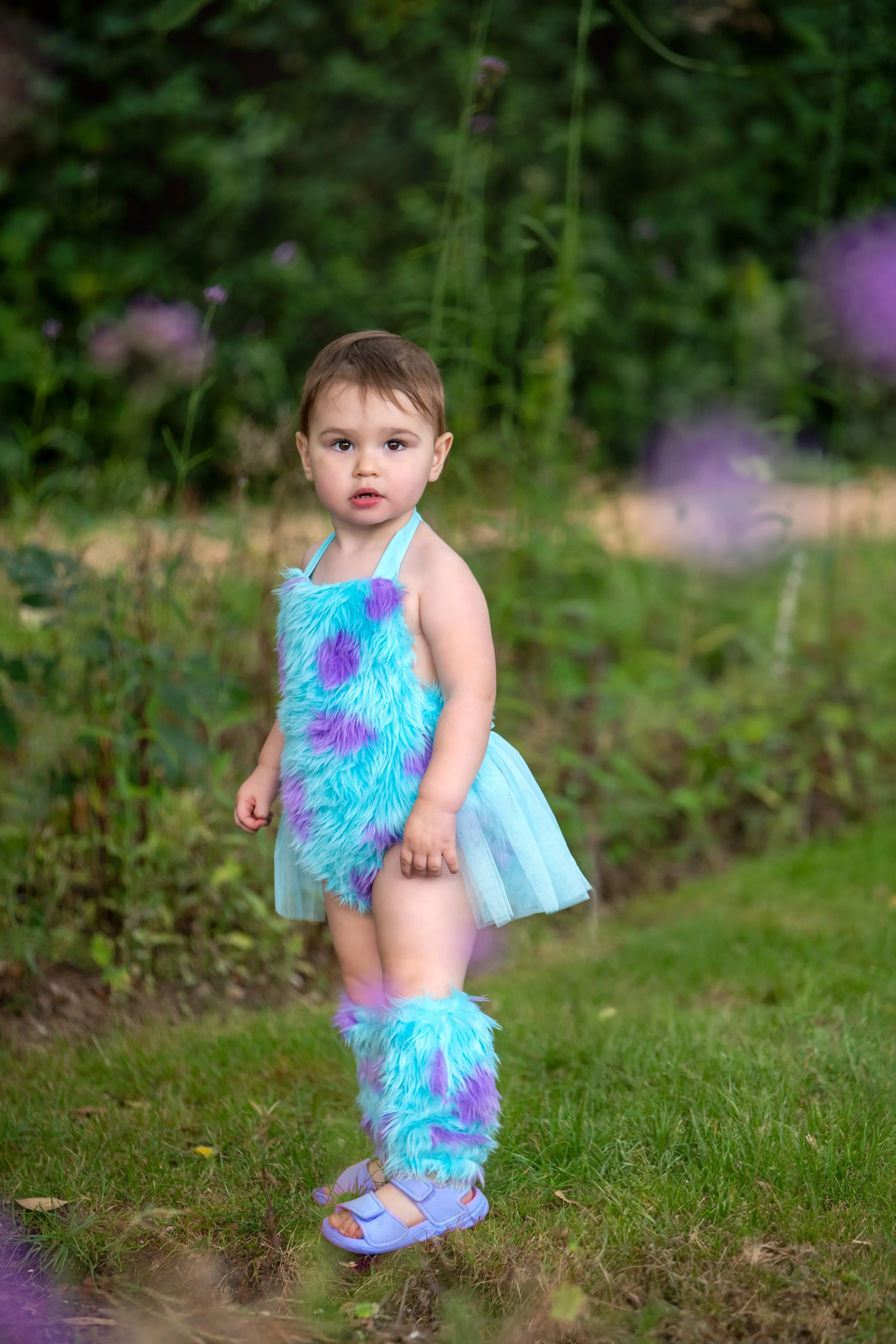 Toddler poses confidently in a fluffy turquoise-and-purple character outfit in a sunlit summer meadow