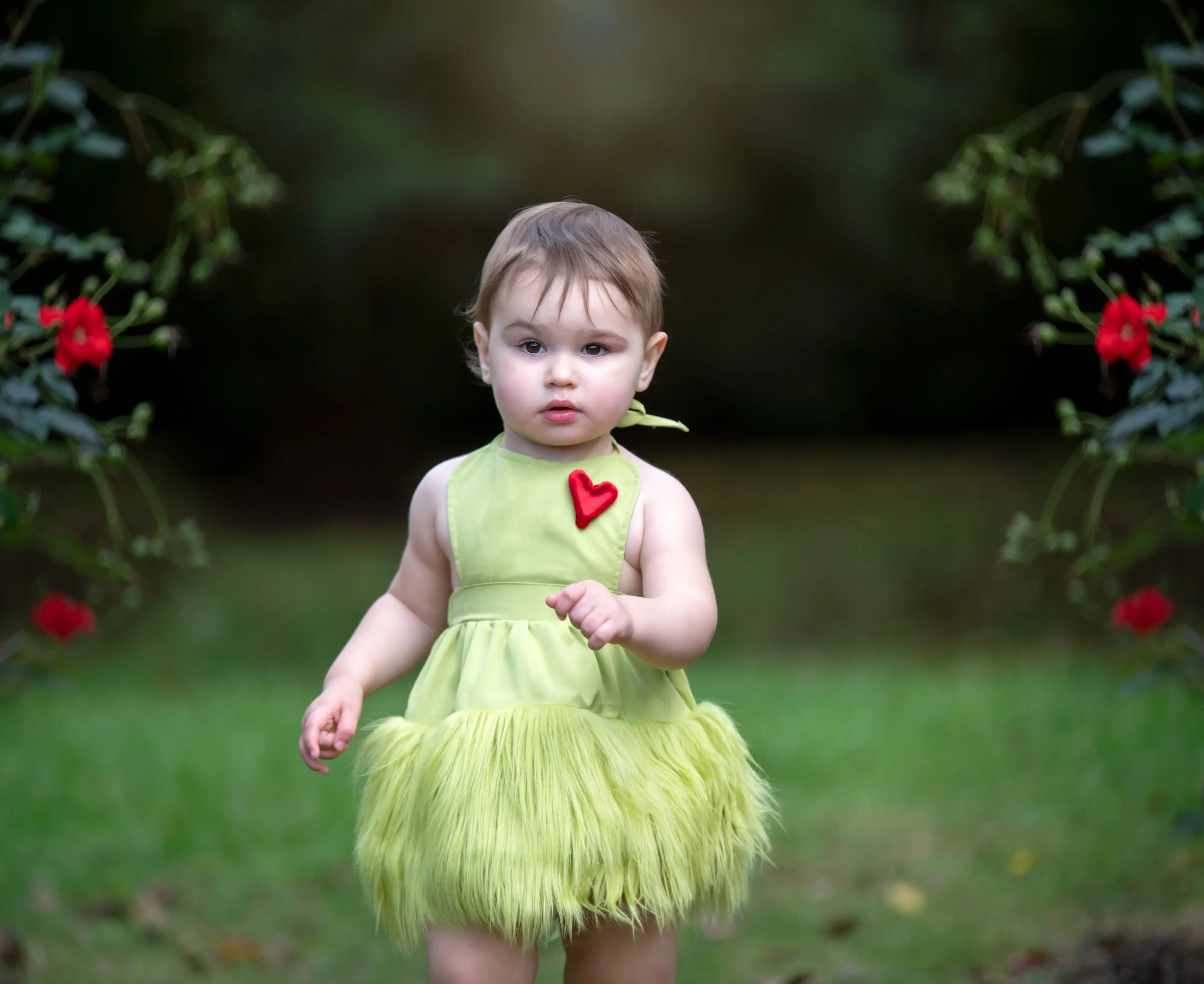 Toddler in green tutu with red heart applique stands between rose arches in soft autumn-garden light