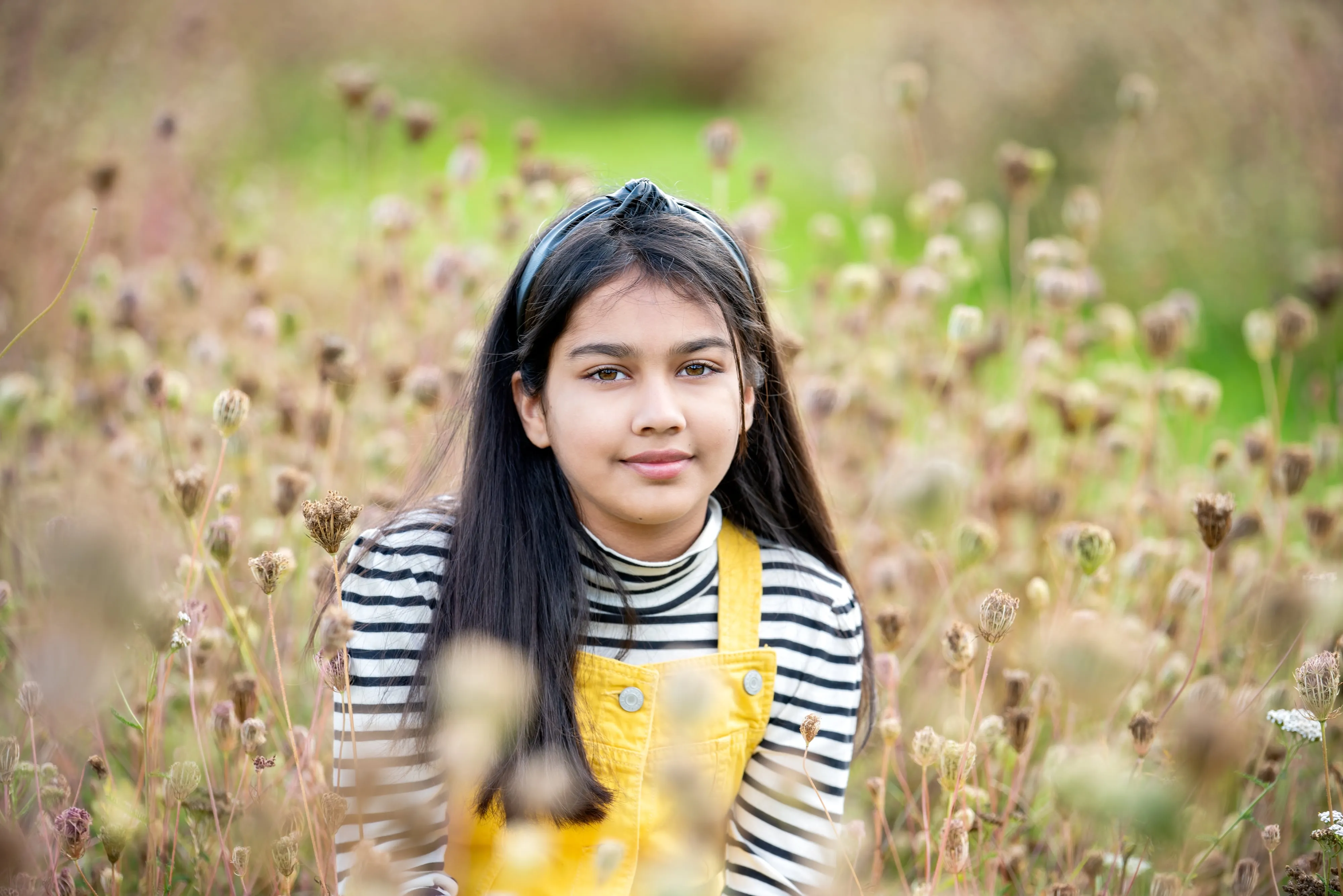 Young girl in yellow corduroy dungarees and striped top crouches in a dried thistle meadow, calm and contemplative