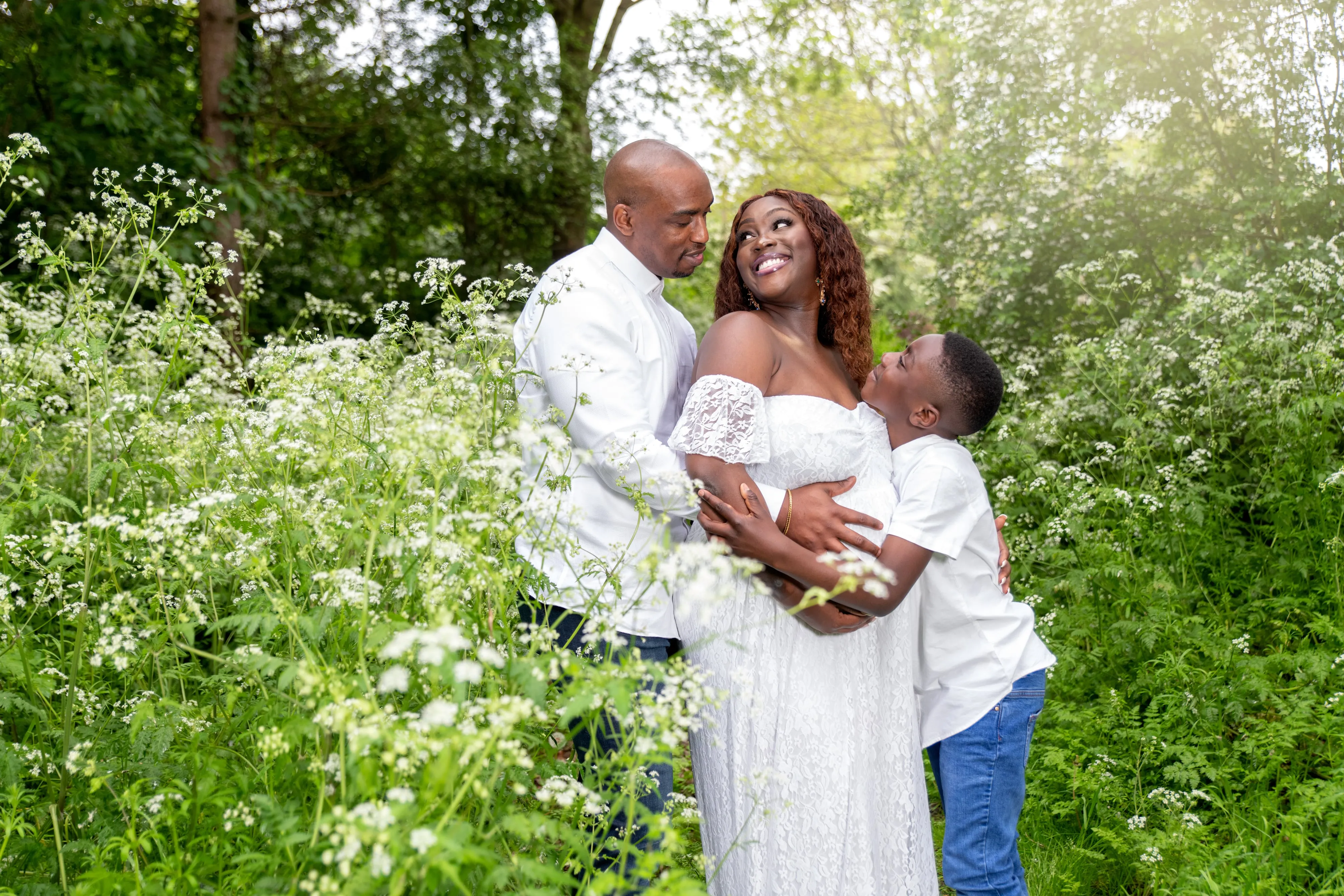 Maternity session in waist-high cow parsley — expecting mum in white lace gown held by her husband and young son