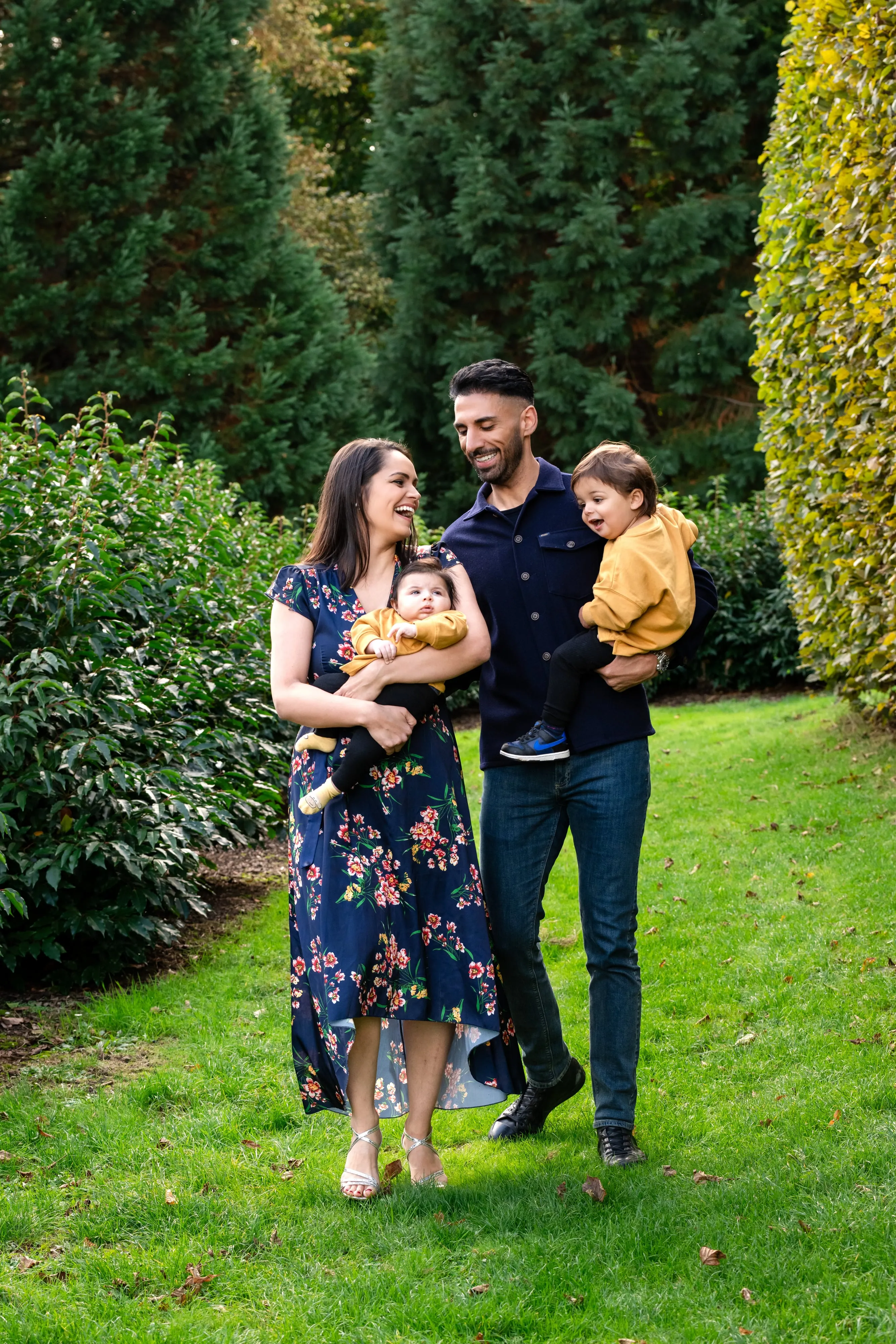 Family of four laugh together on the lawn at Willen Lake — mum in floral dress holding baby in mustard, dad with toddler in his arms — surrounded by autumn topiary