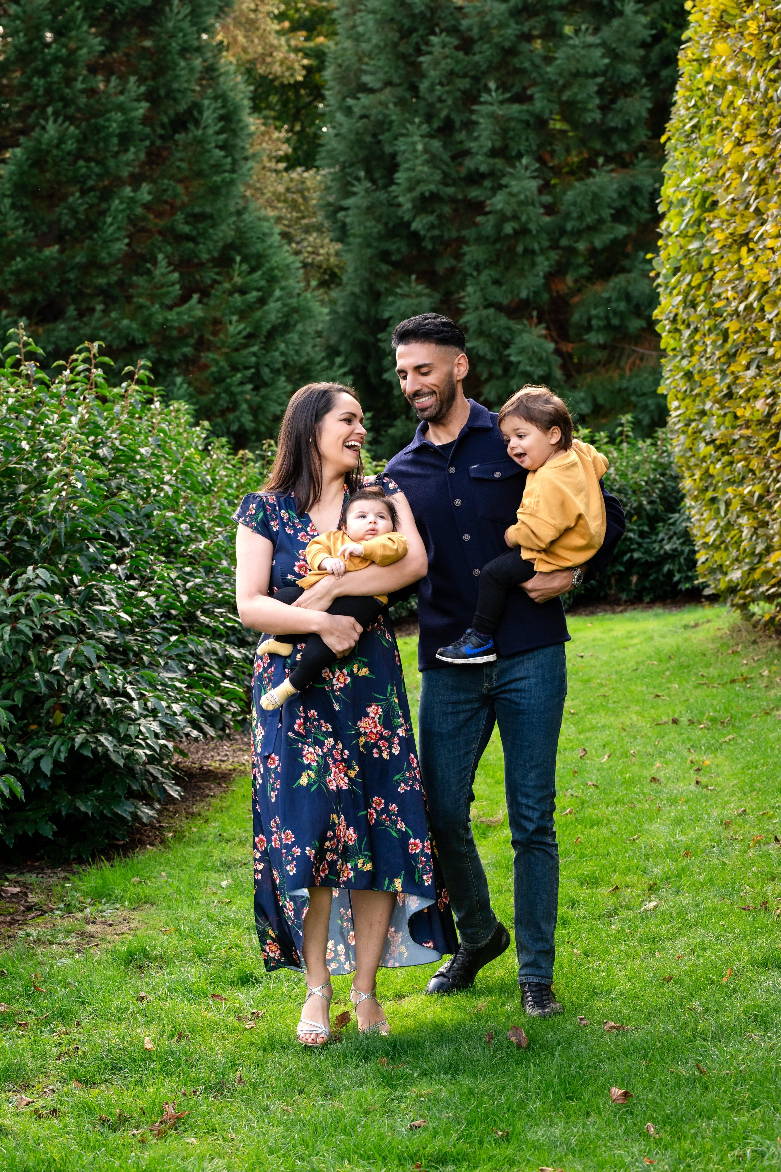 Family of four laugh together on the lawn at Willen Lake — mum in floral dress holding baby in mustard, dad with toddler in his arms — surrounded by autumn topiary