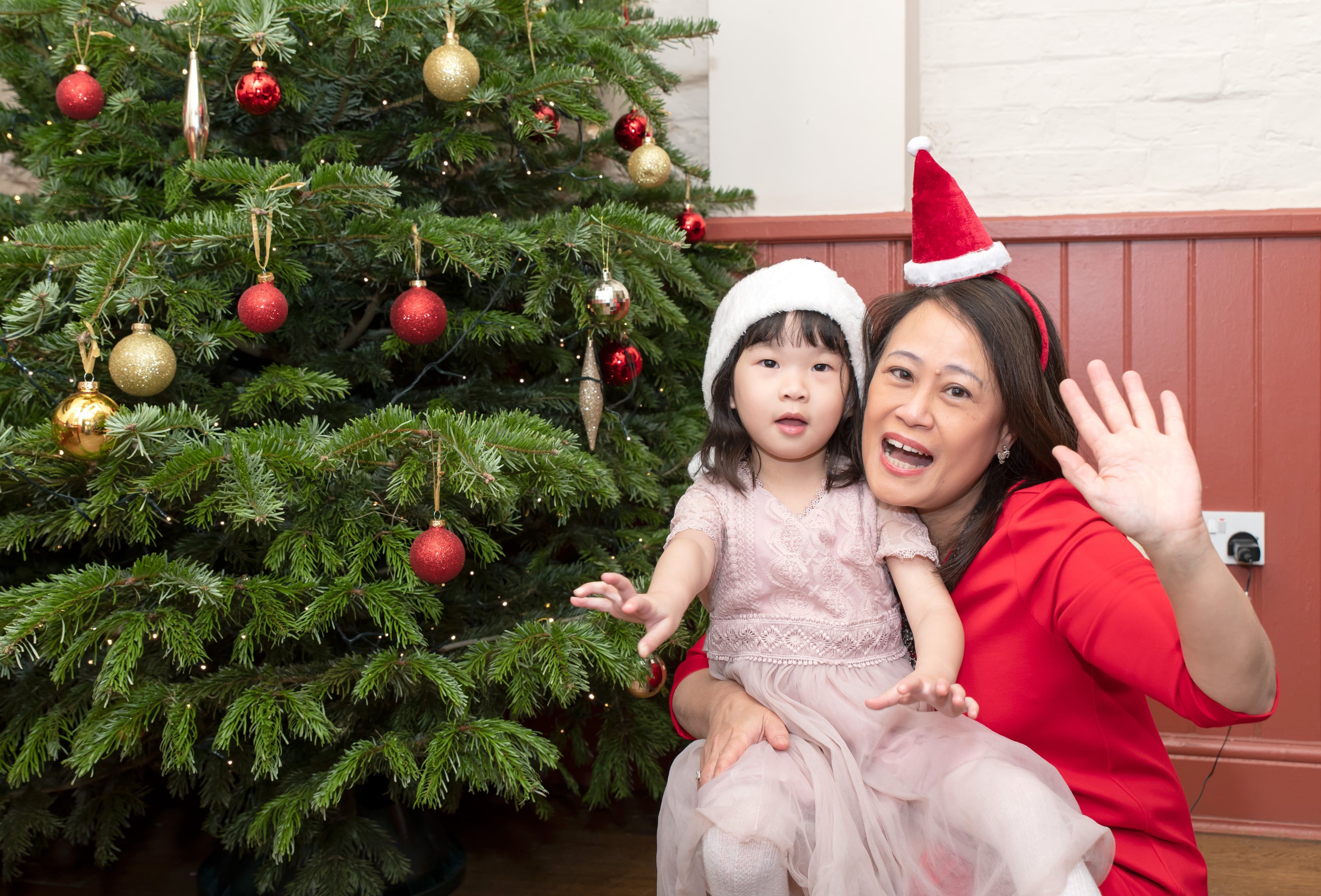 Mother in a Santa hat and her little girl wave and laugh together beside the Christmas tree