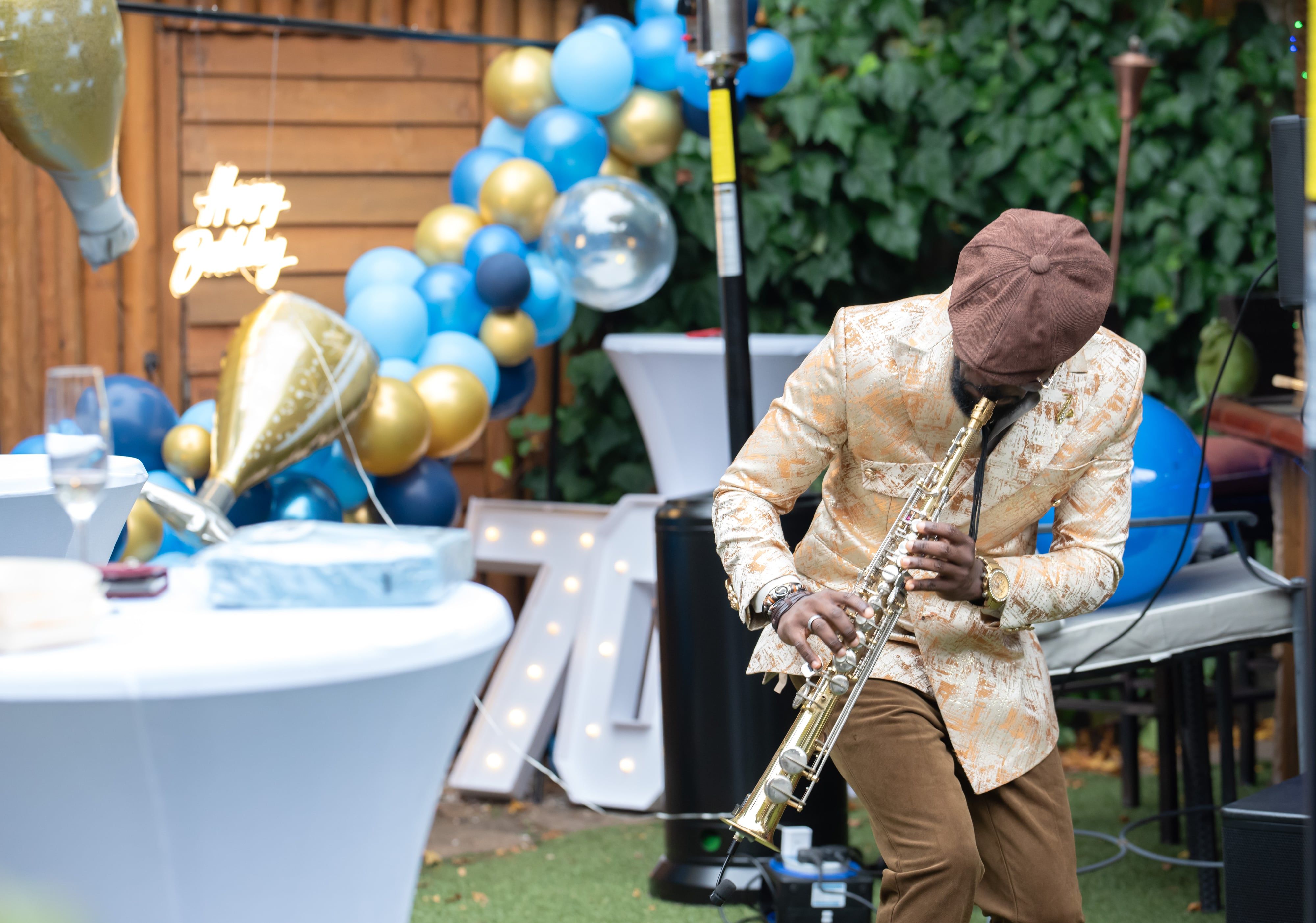 Saxophonist performs beside a blue-and-gold balloon arch at an outdoor birthday party