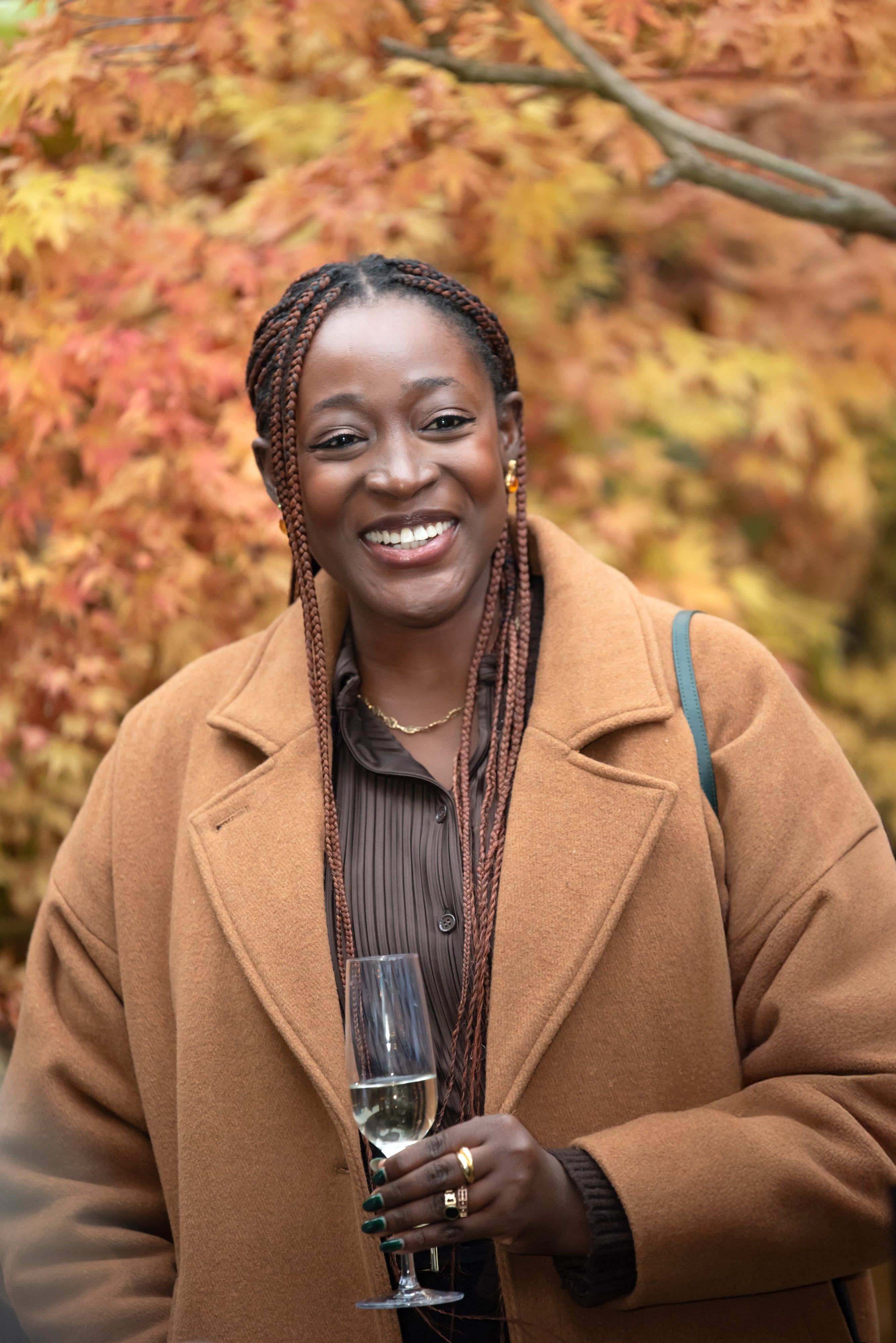 Smiling guest in a camel coat raises a glass of fizz before vivid autumn maple leaves