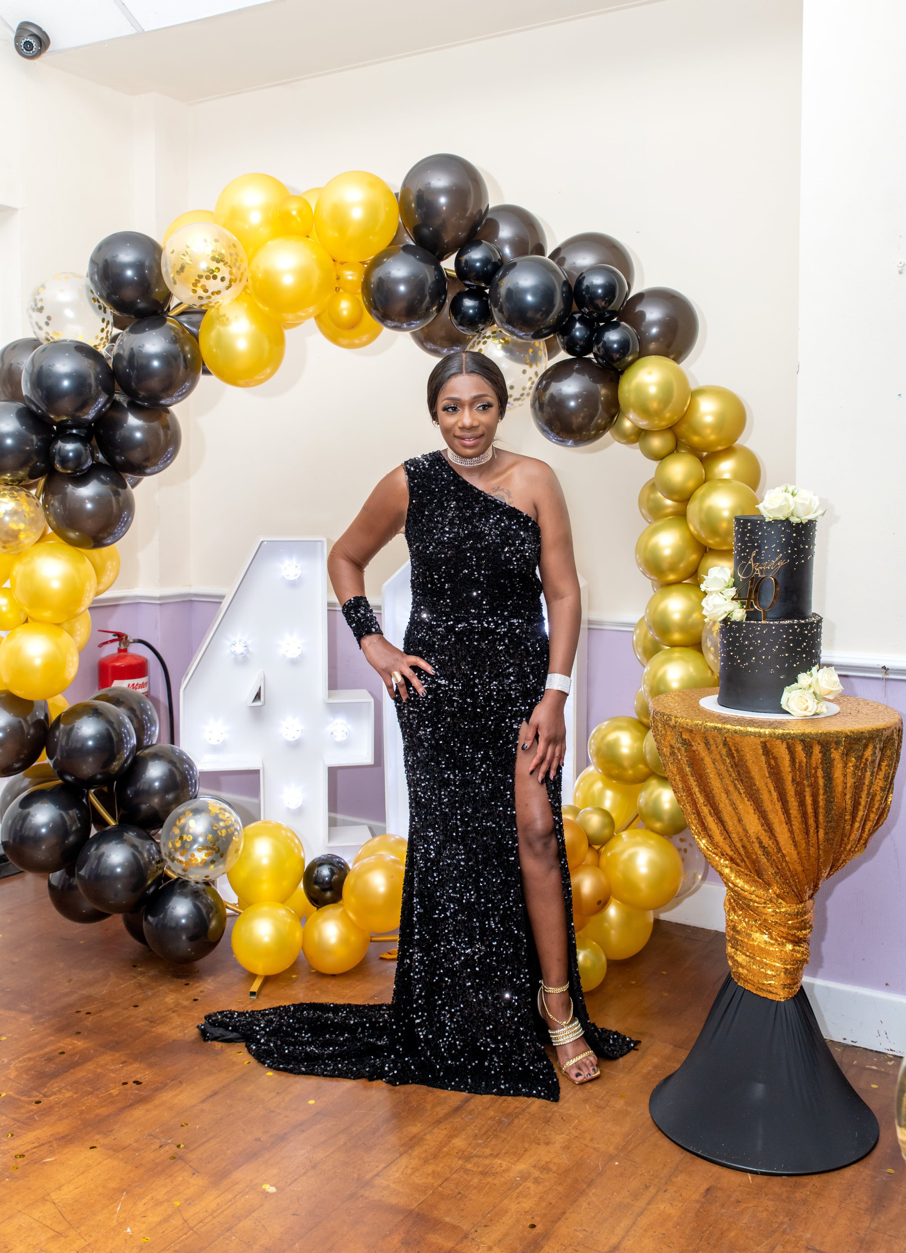 Guest in a sequinned black gown beside a black-and-gold balloon arch and tiered cake