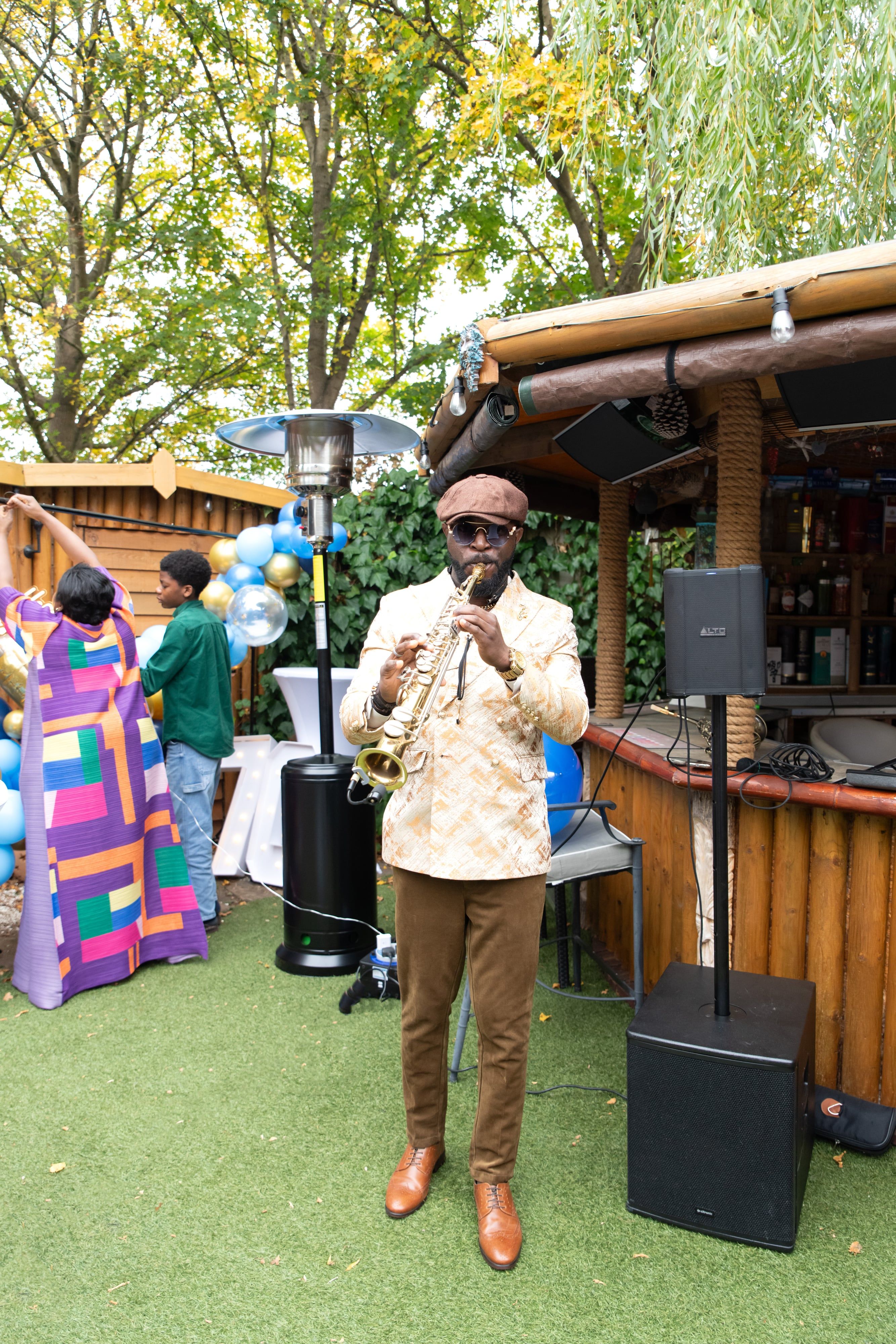 Saxophonist in a cream jacket performs for guests at a balloon-decked garden party