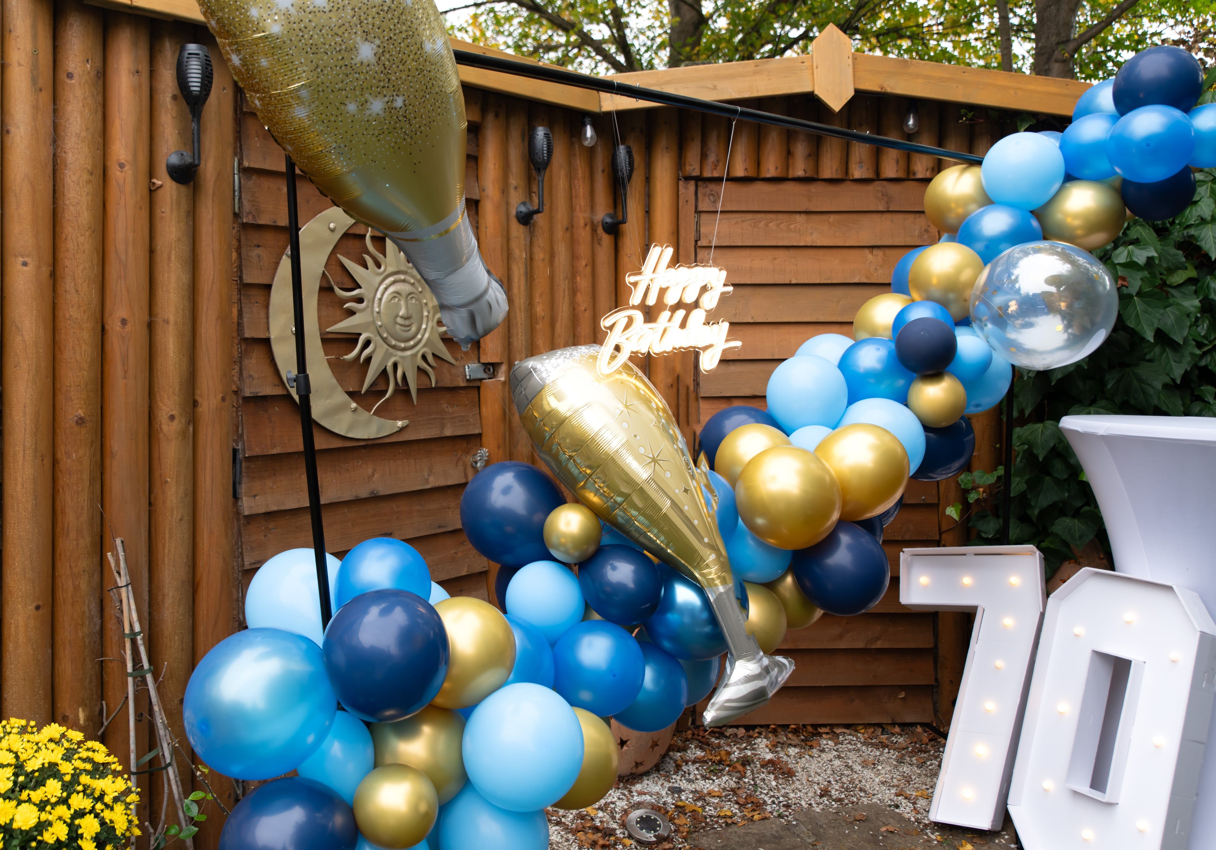 Navy, blue and gold balloon arch with champagne-bottle balloons and a light-up 70 at a garden party