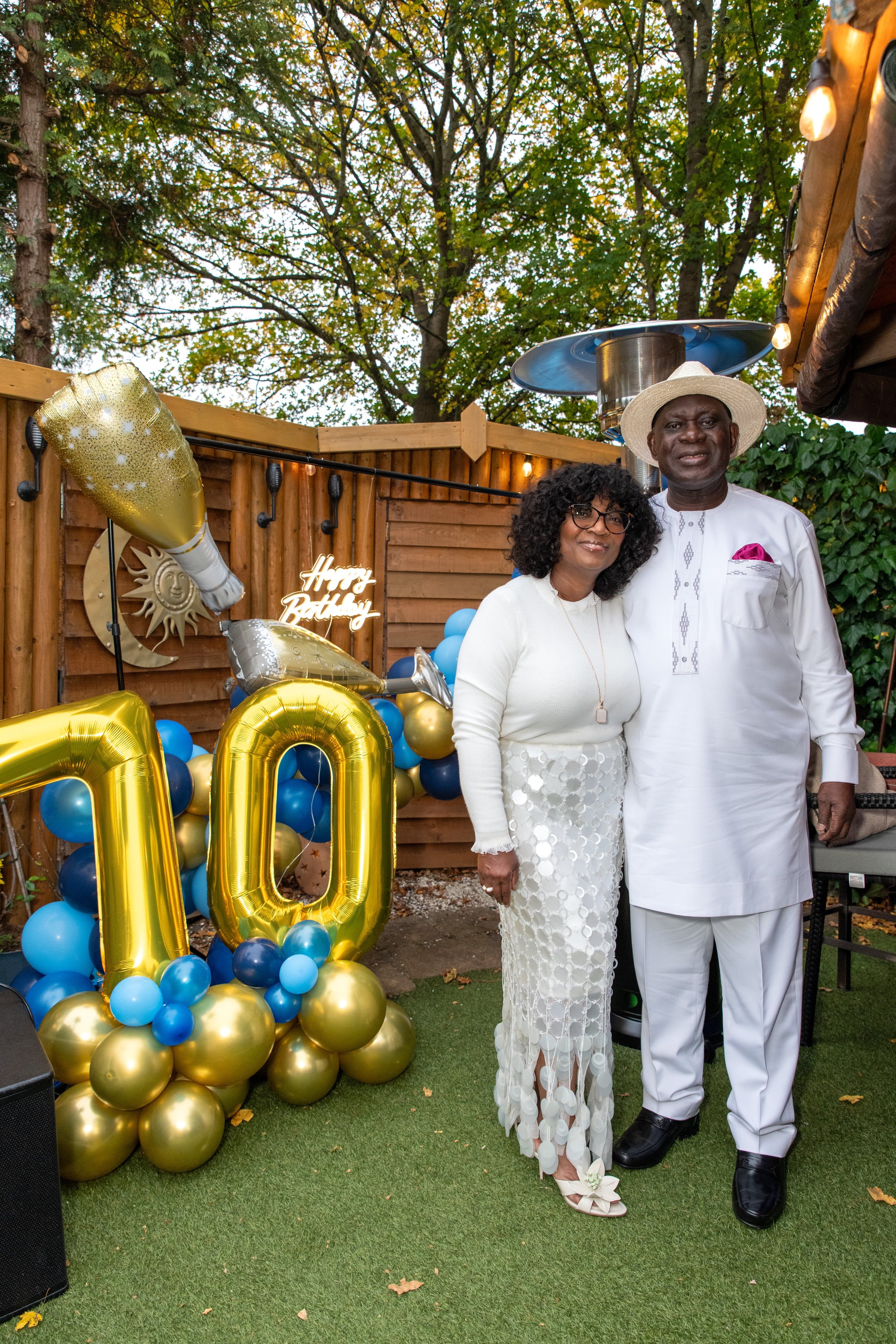 Couple in coordinating white outfits beside the gold 70 balloons at their garden party