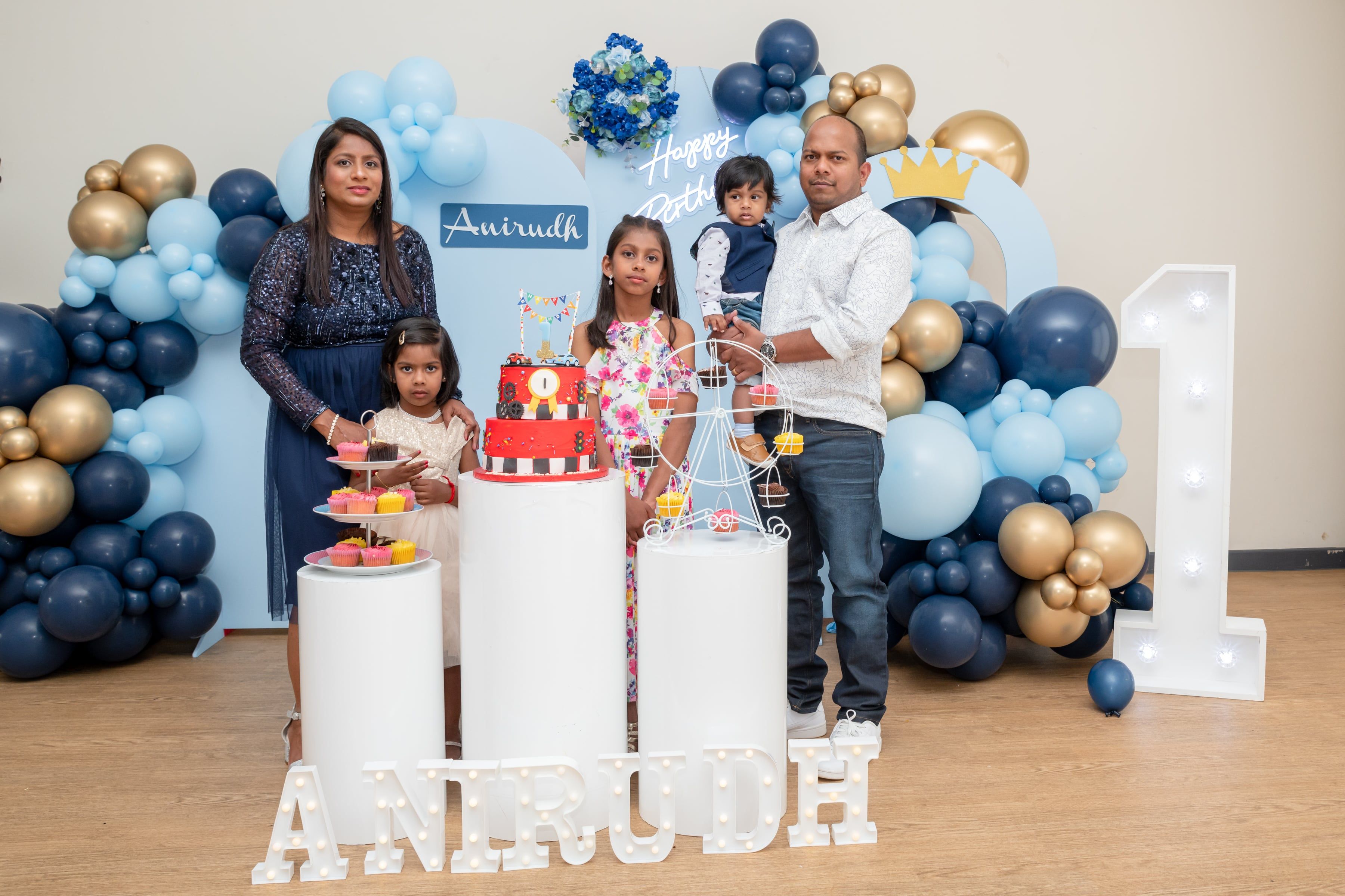 Family of five together at a blue-and-gold first-birthday backdrop with a light-up number one