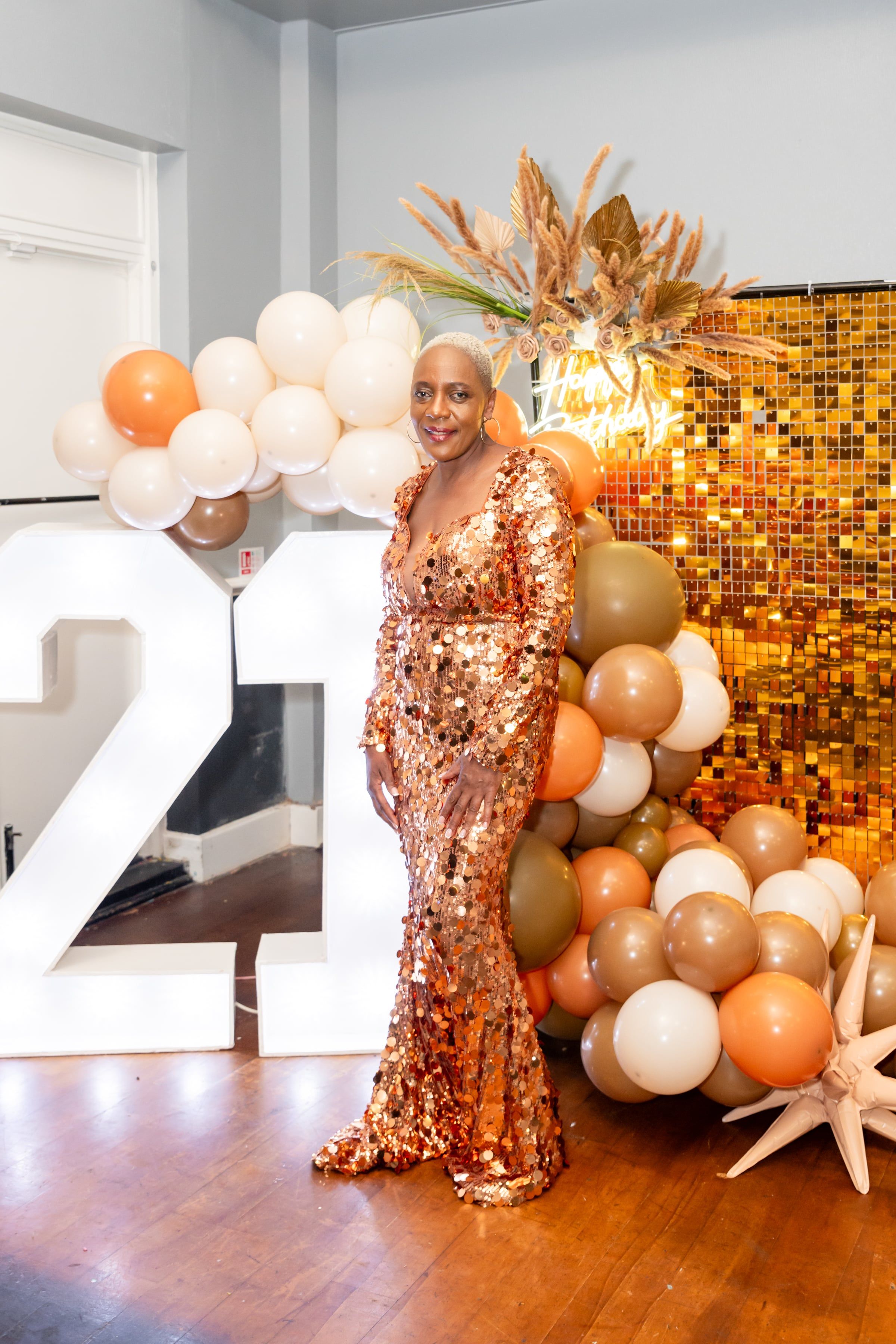 Guest in a copper sequinned gown before a shimmering gold wall and balloon arch at an evening party