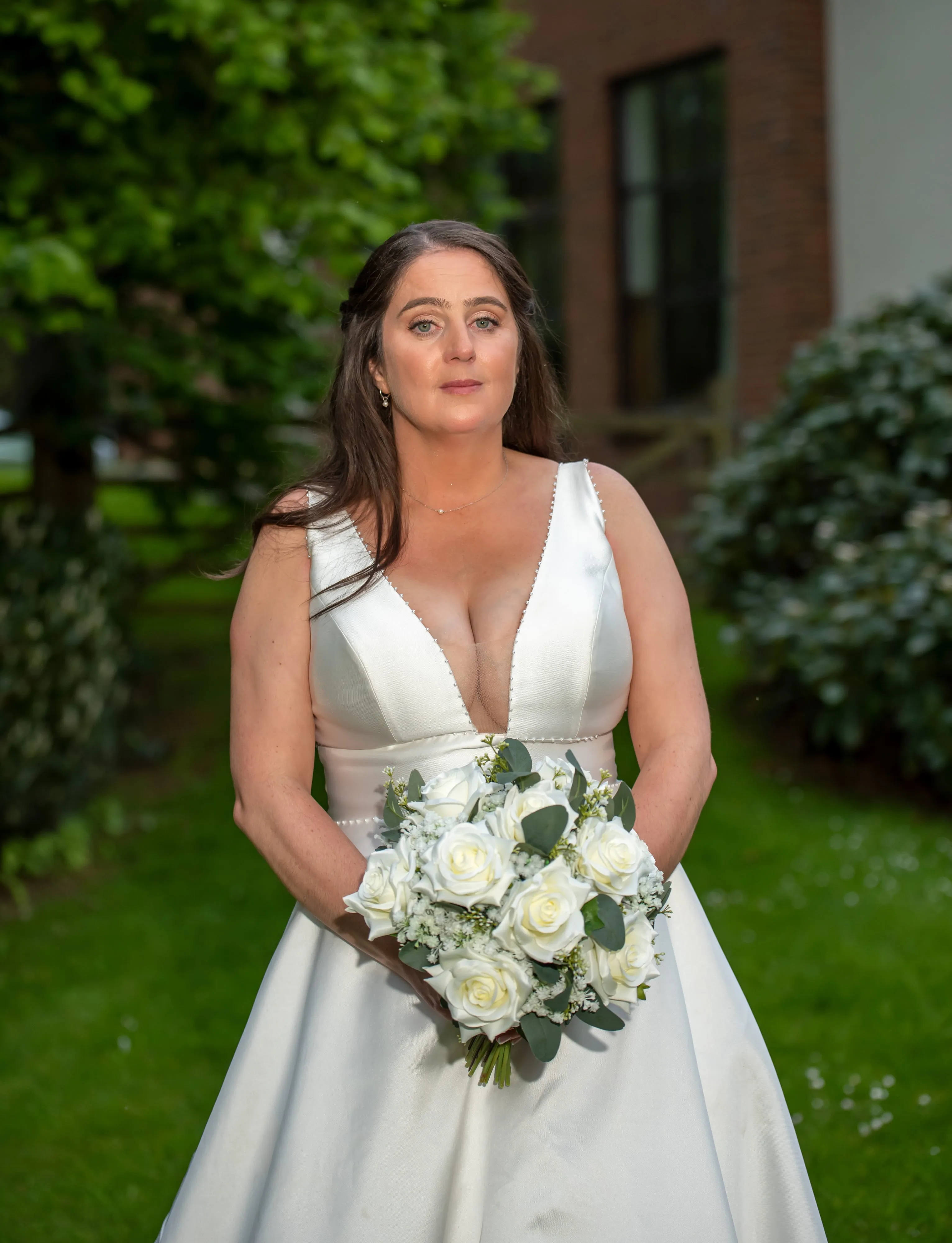 Solo bridal portrait — bride in white satin v-neck gown holding a tight white-rose bouquet against the Belfry gardens