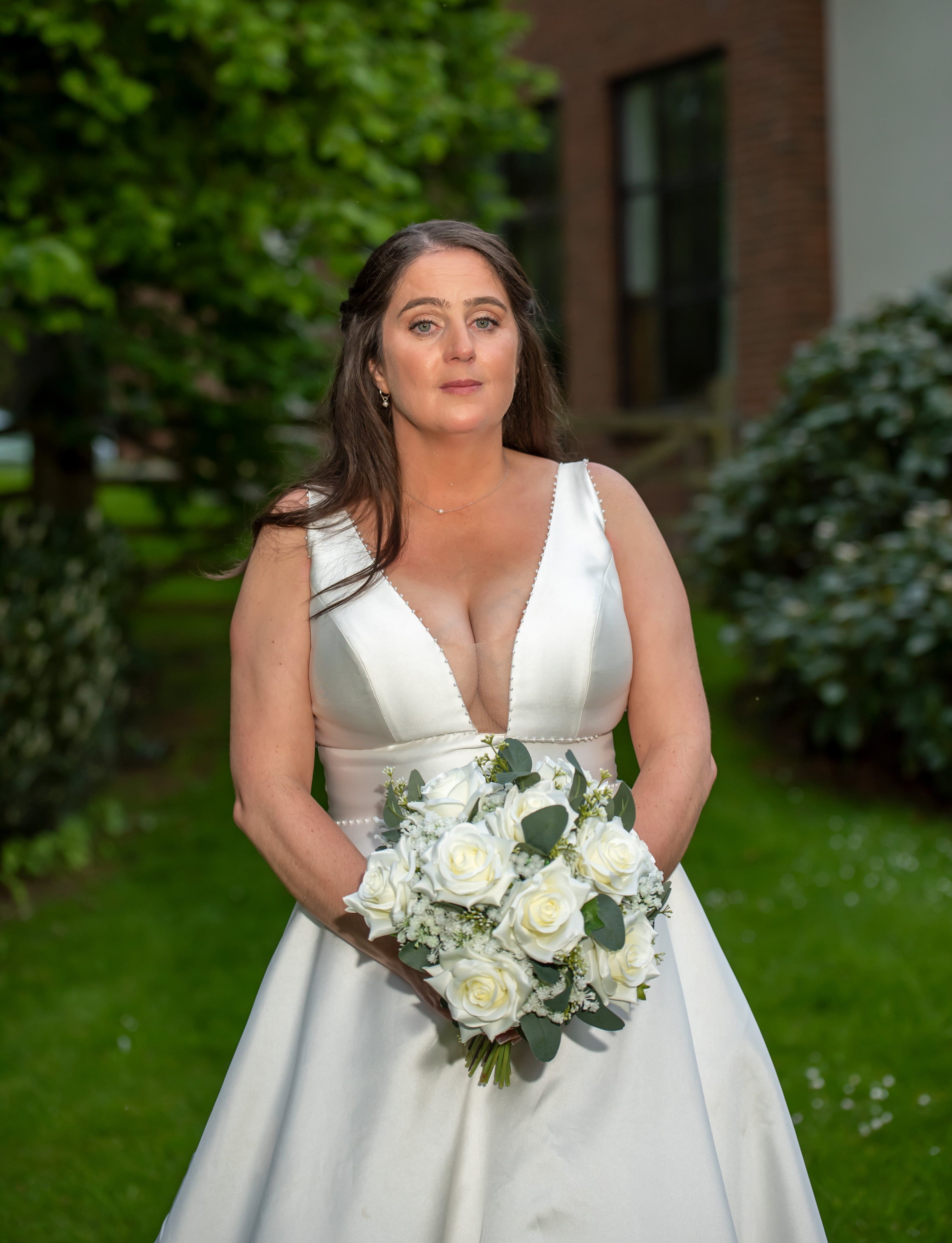 Solo bridal portrait — bride in white satin v-neck gown holding a tight white-rose bouquet against the Belfry gardens