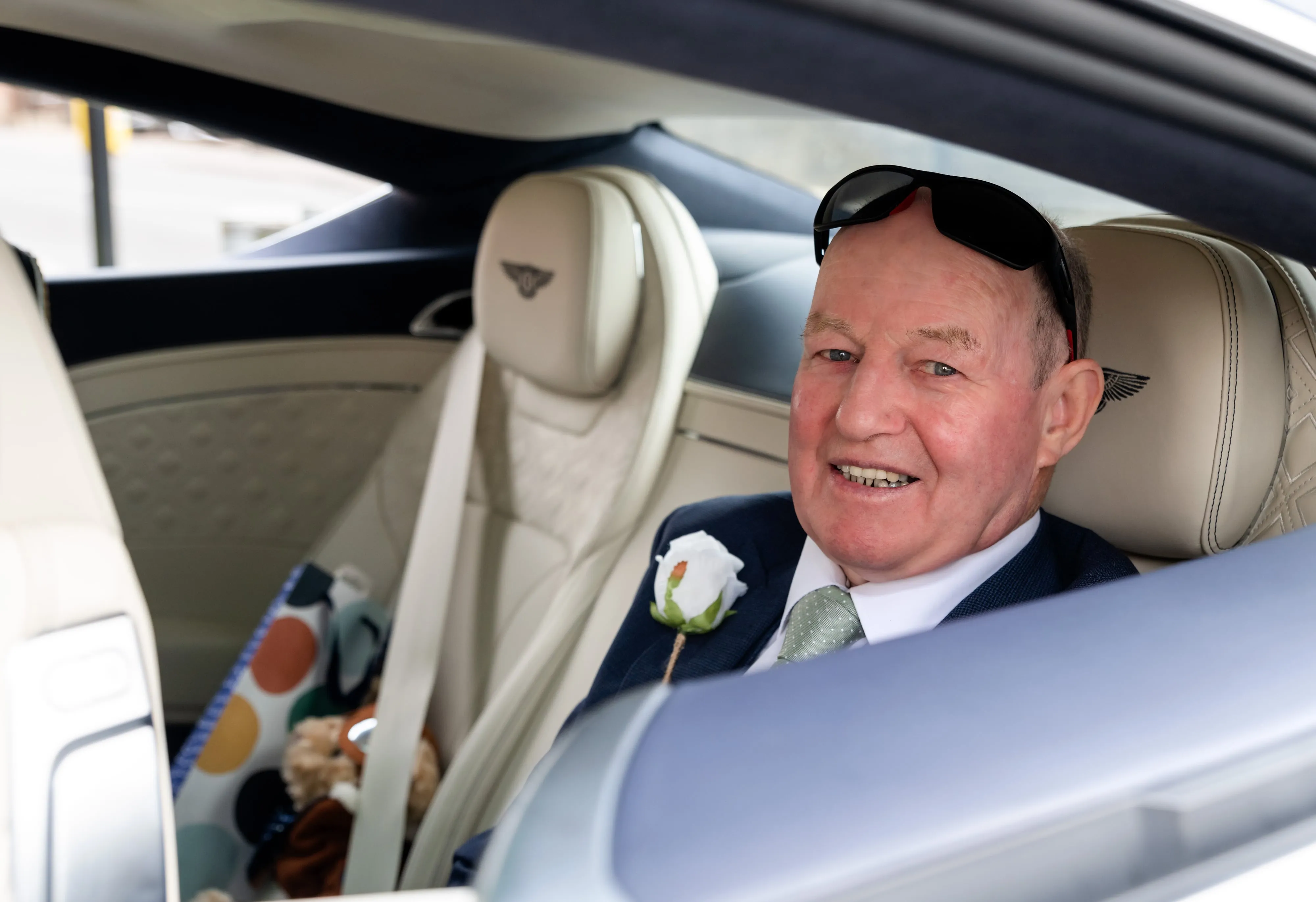 Father of the bride relaxes in the Bentley with a white rose buttonhole, sunglasses on, post-arrival candid