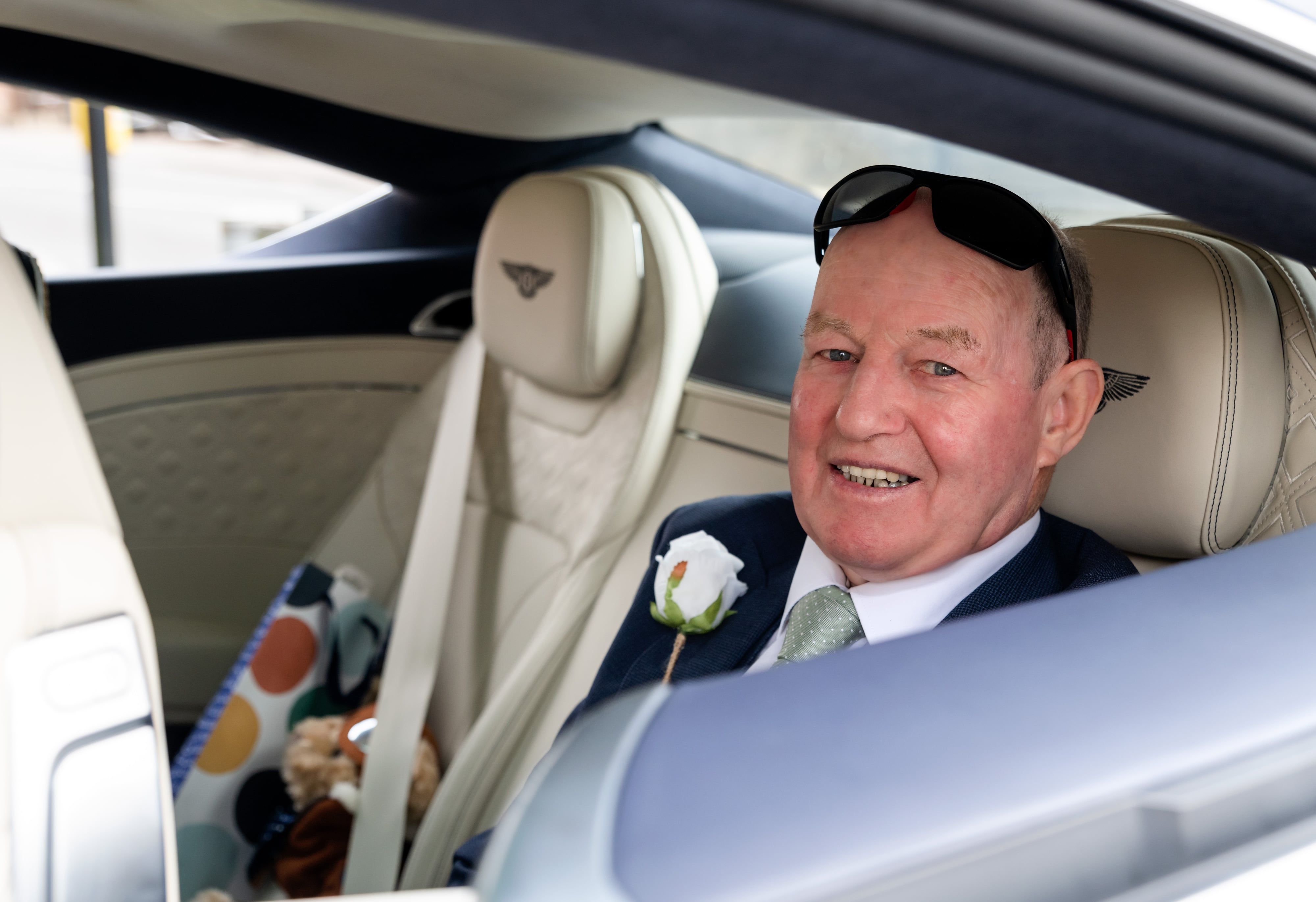 Father of the bride relaxes in the Bentley with a white rose buttonhole, sunglasses on, post-arrival candid