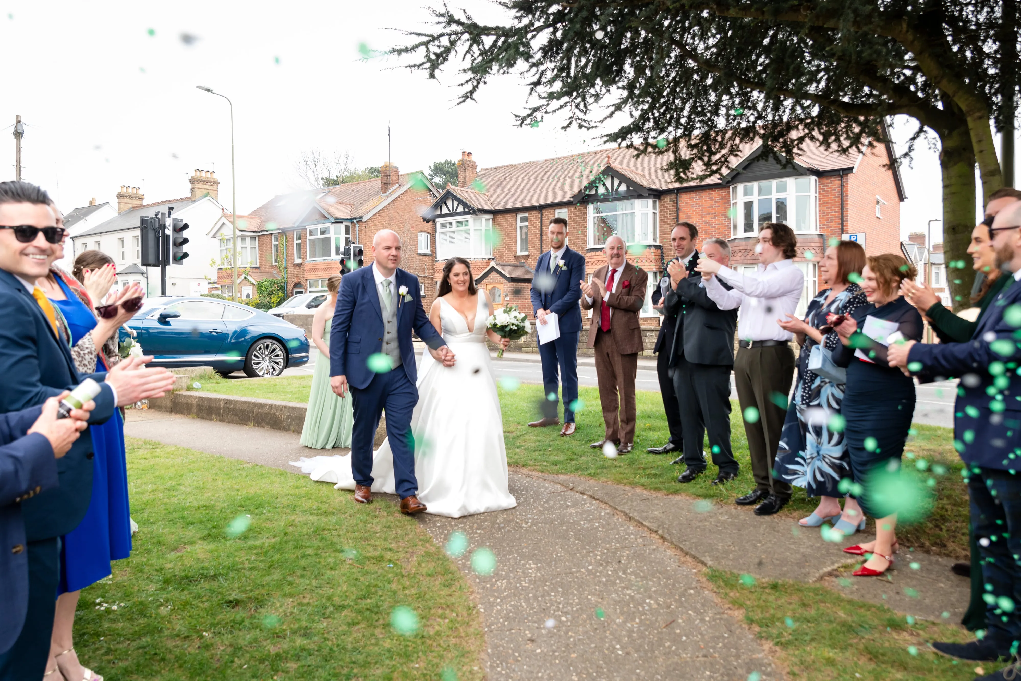 Bride and groom walk down confetti aisle with cheering guests outside venue