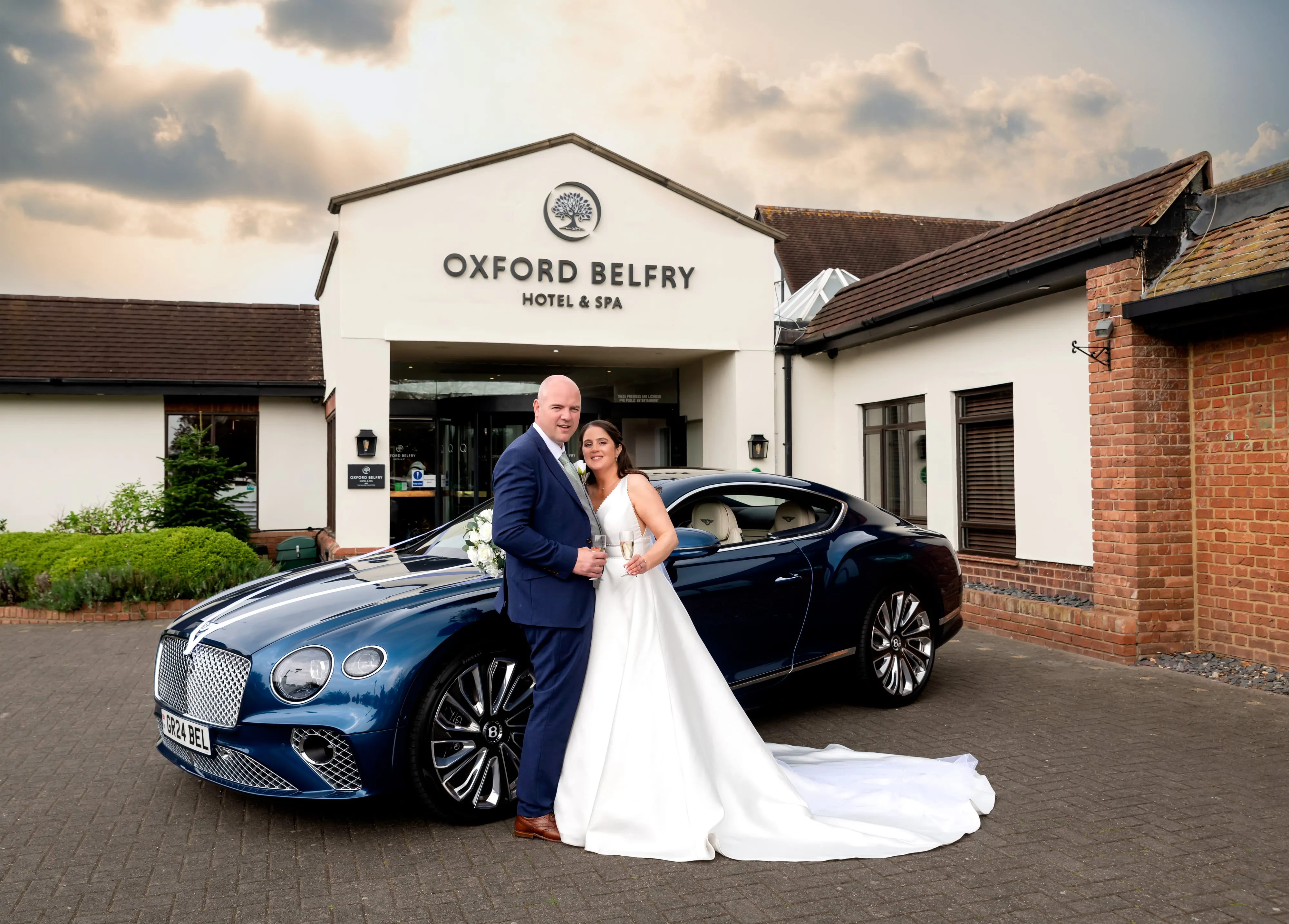 Bride and groom pose with blue Bentley outside Oxford Belfry Hotel and Spa entrance