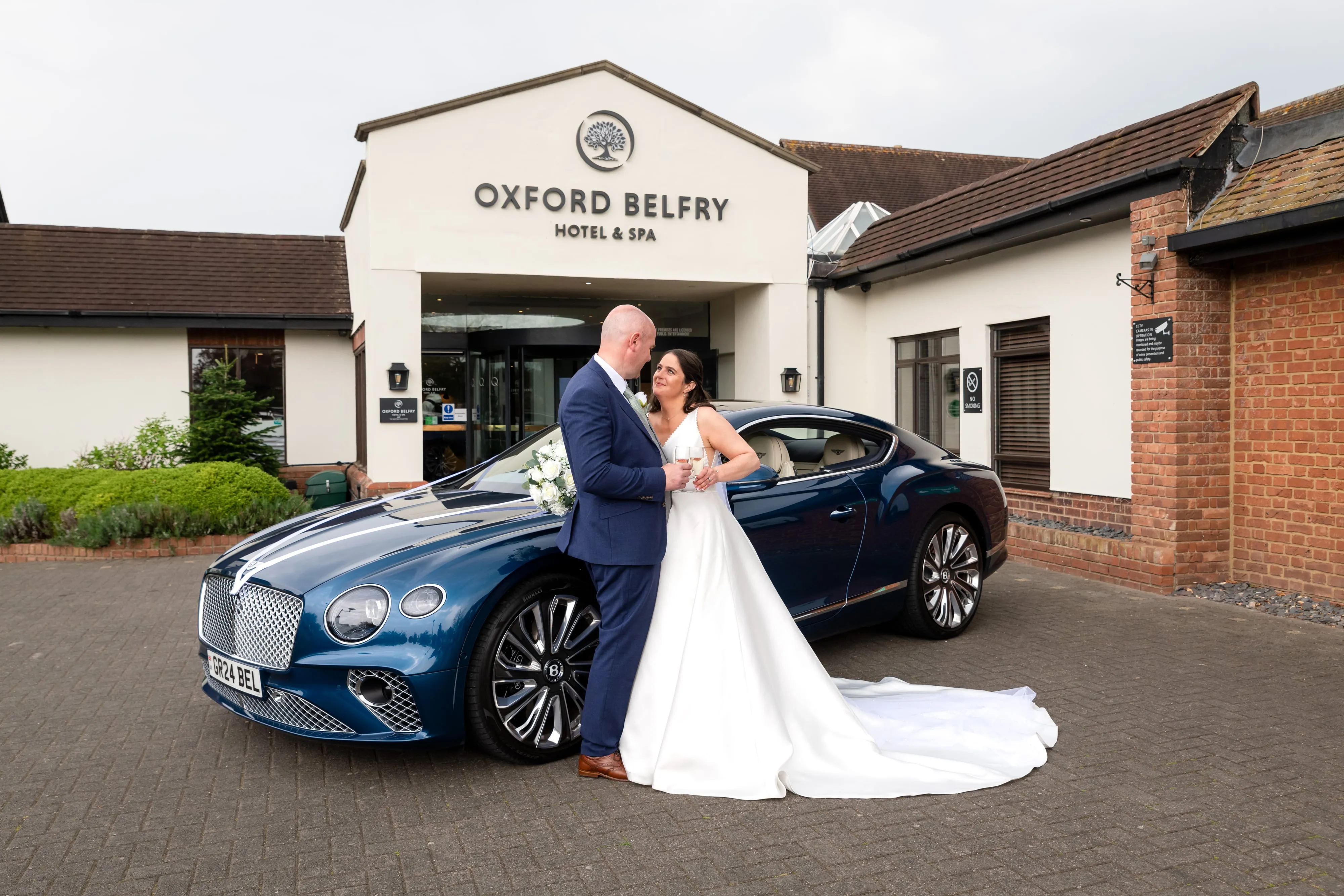 Bride and groom share an intimate moment beside a blue Bentley GT under the Oxford Belfry signed entrance