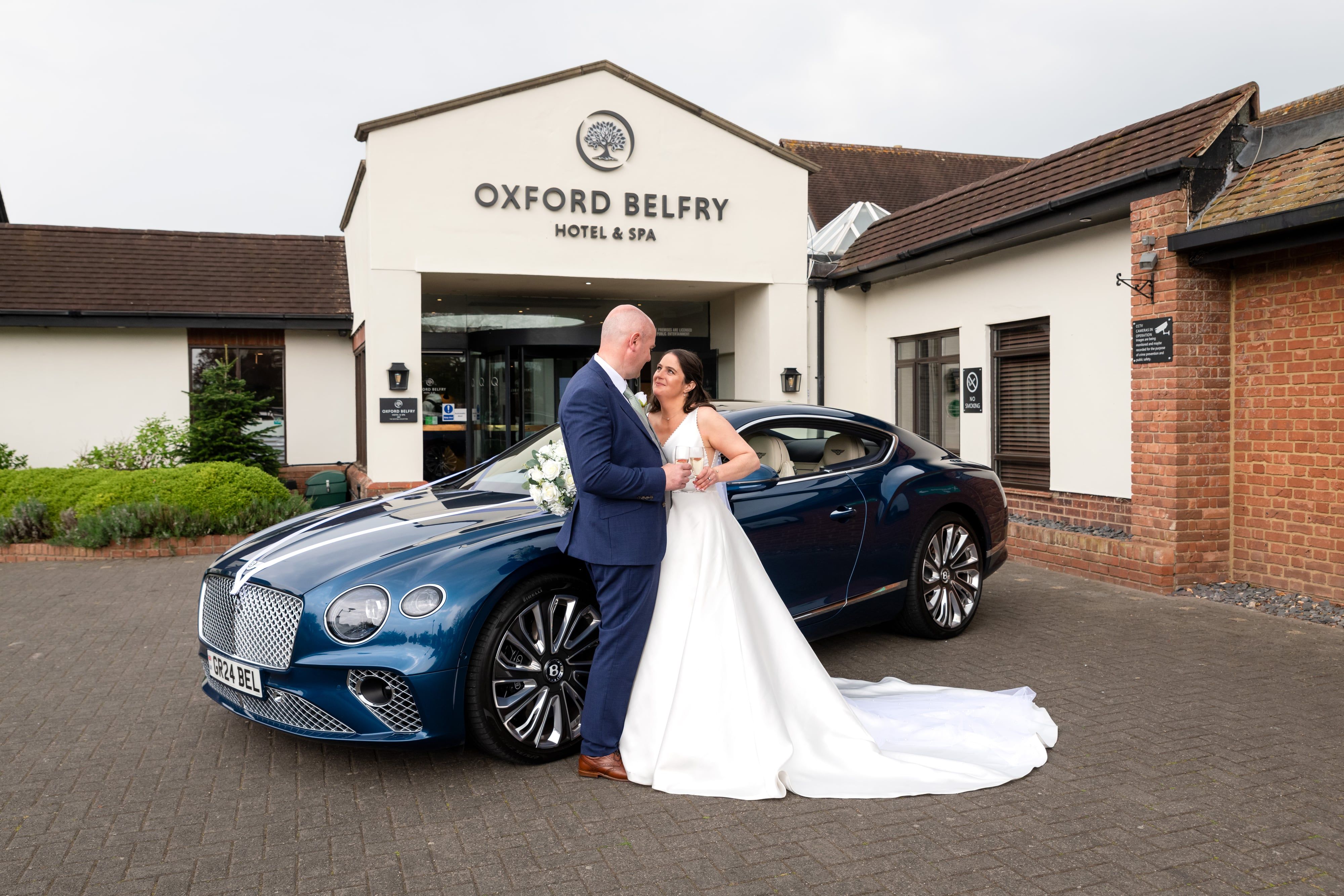Bride and groom share an intimate moment beside a blue Bentley GT under the Oxford Belfry signed entrance
