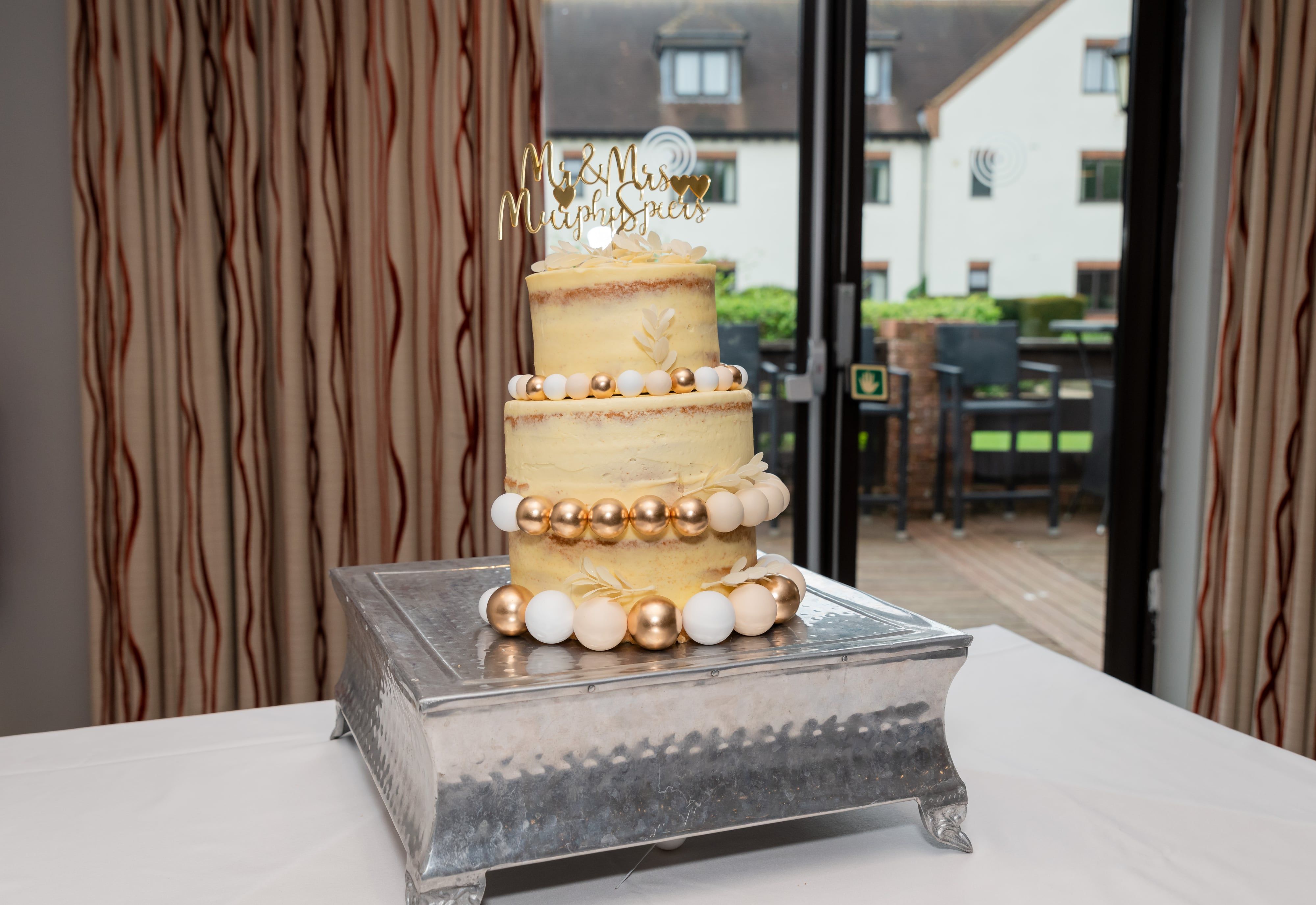 Two-tier wedding cake with Mr & Mrs Murphy-Spiers gold topper and gold-and-cream beading detail, courtyard view through the window