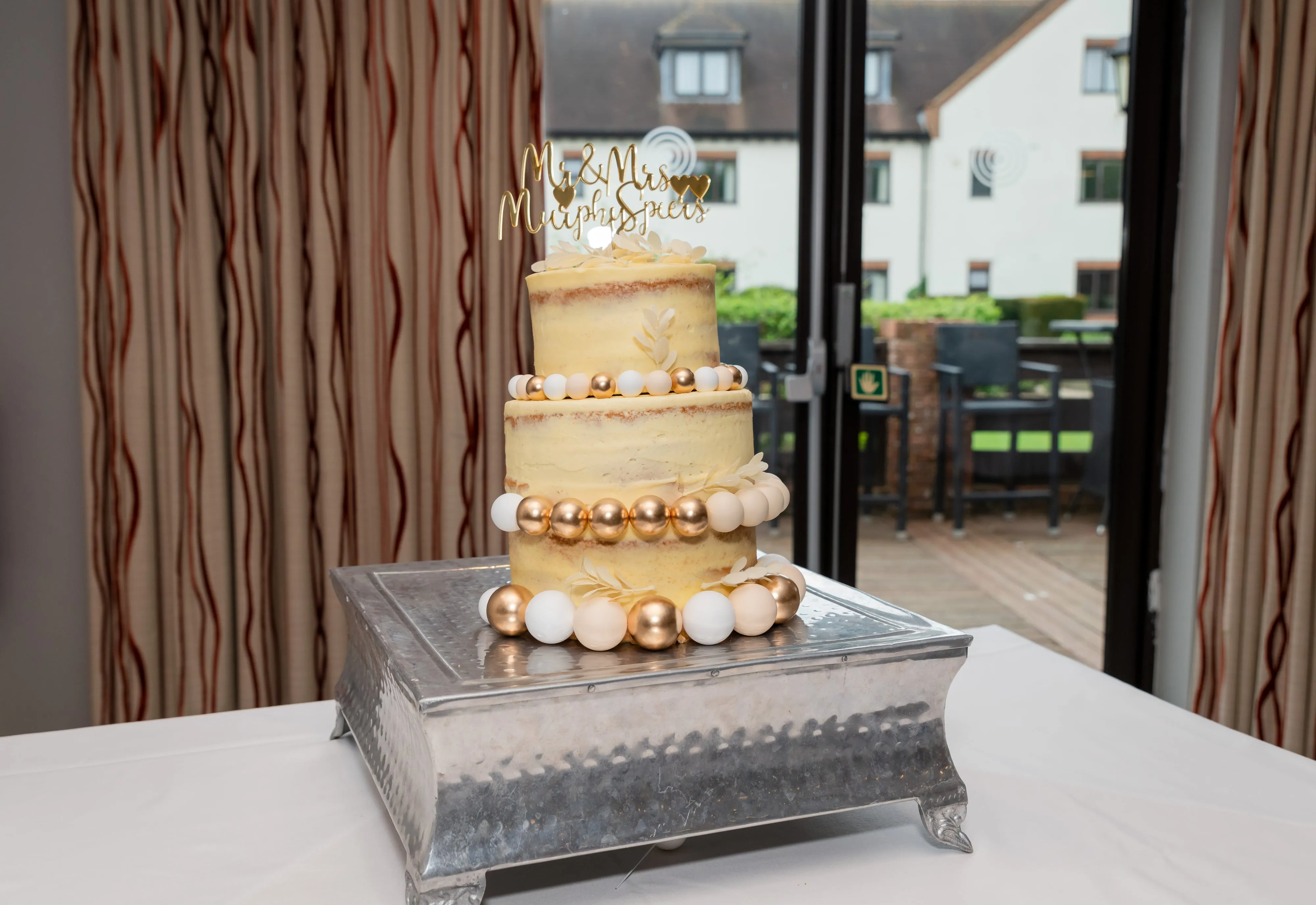 Two-tier wedding cake with Mr & Mrs Murphy-Spiers gold topper and gold-and-cream beading detail, courtyard view through the window