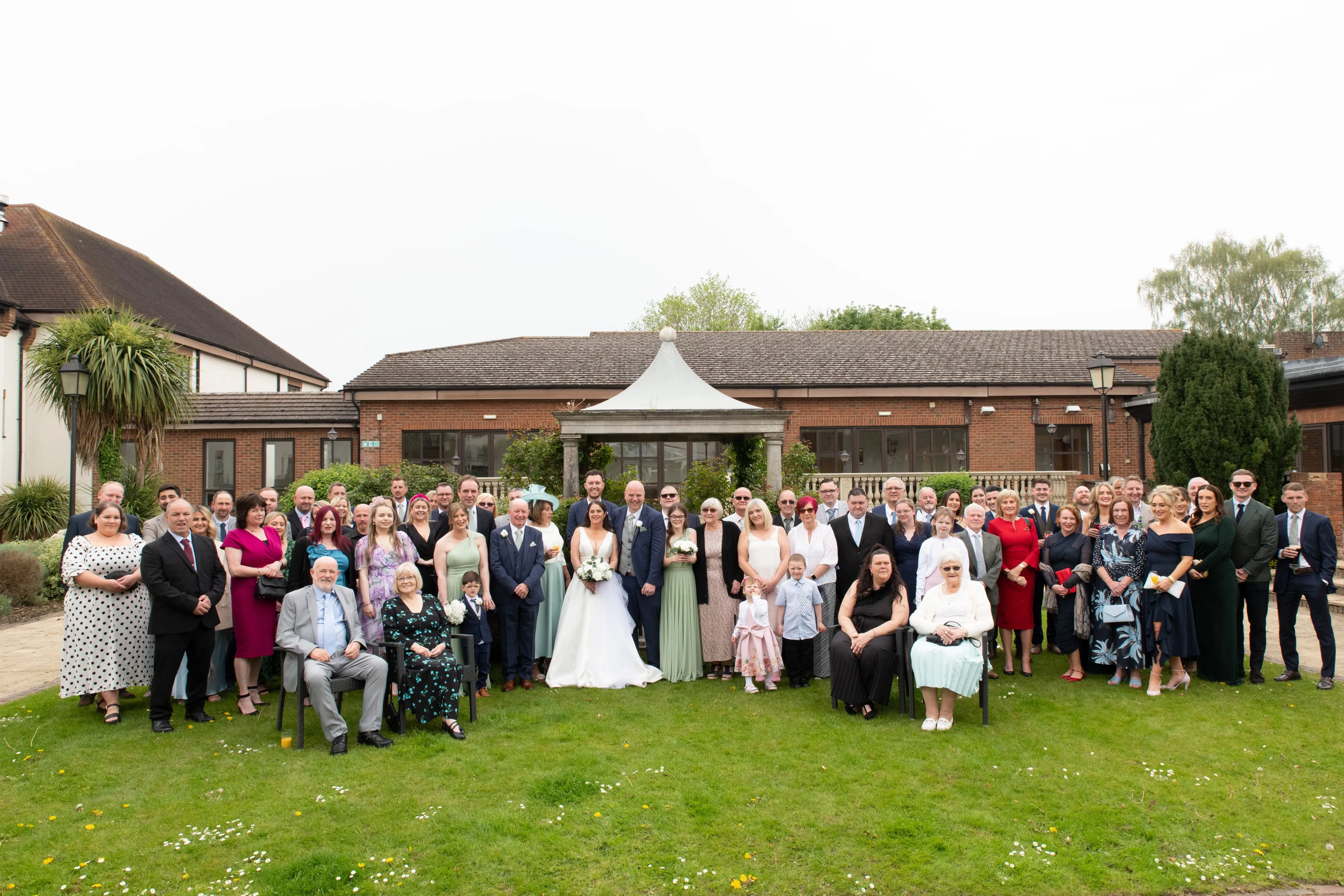 Wide whole-wedding group portrait of fifty-plus guests on the lawn in front of the Belfry stone pavilion