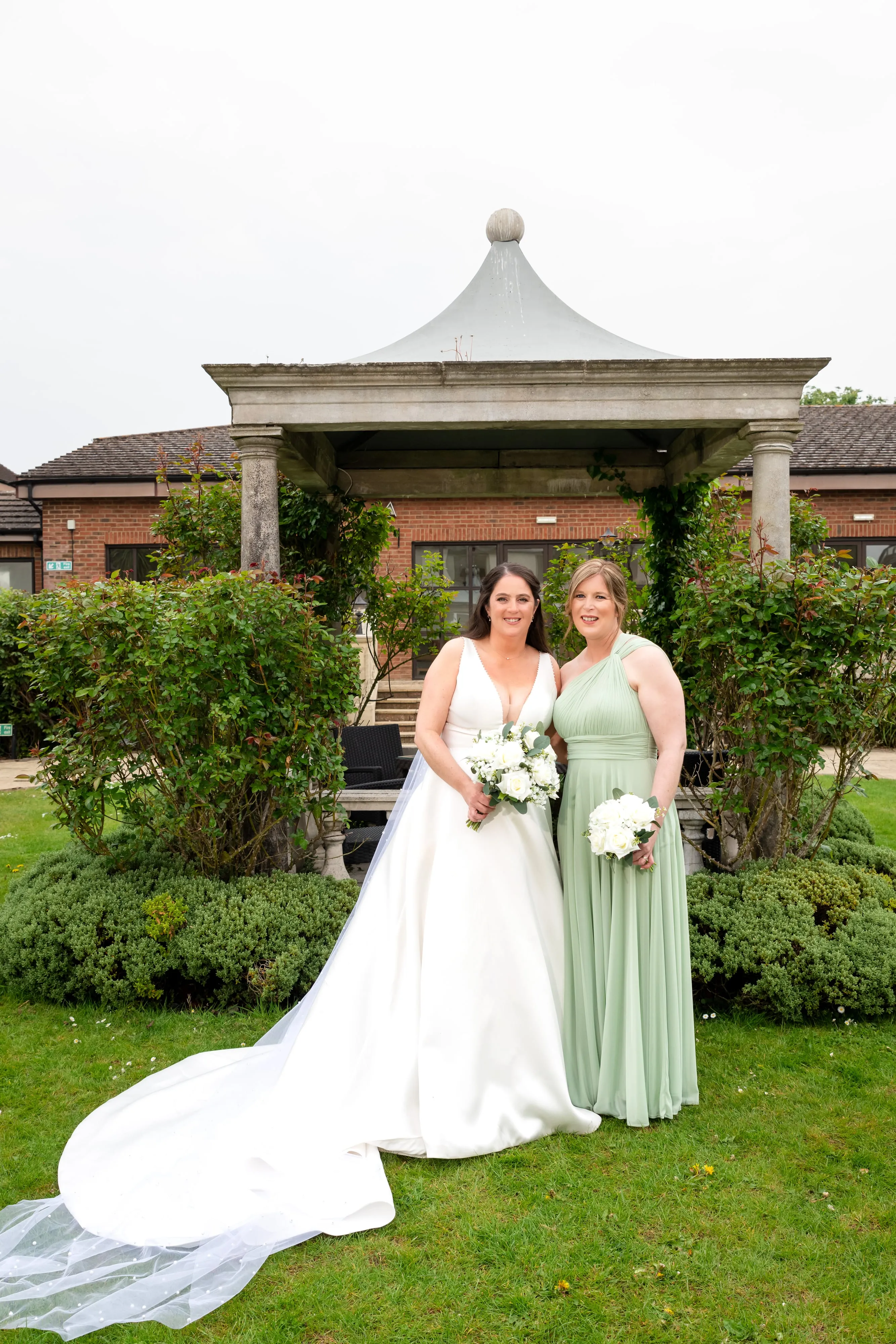 Bride and bridesmaid in sage-green gown stand in front of the stone pavilion at the Belfry