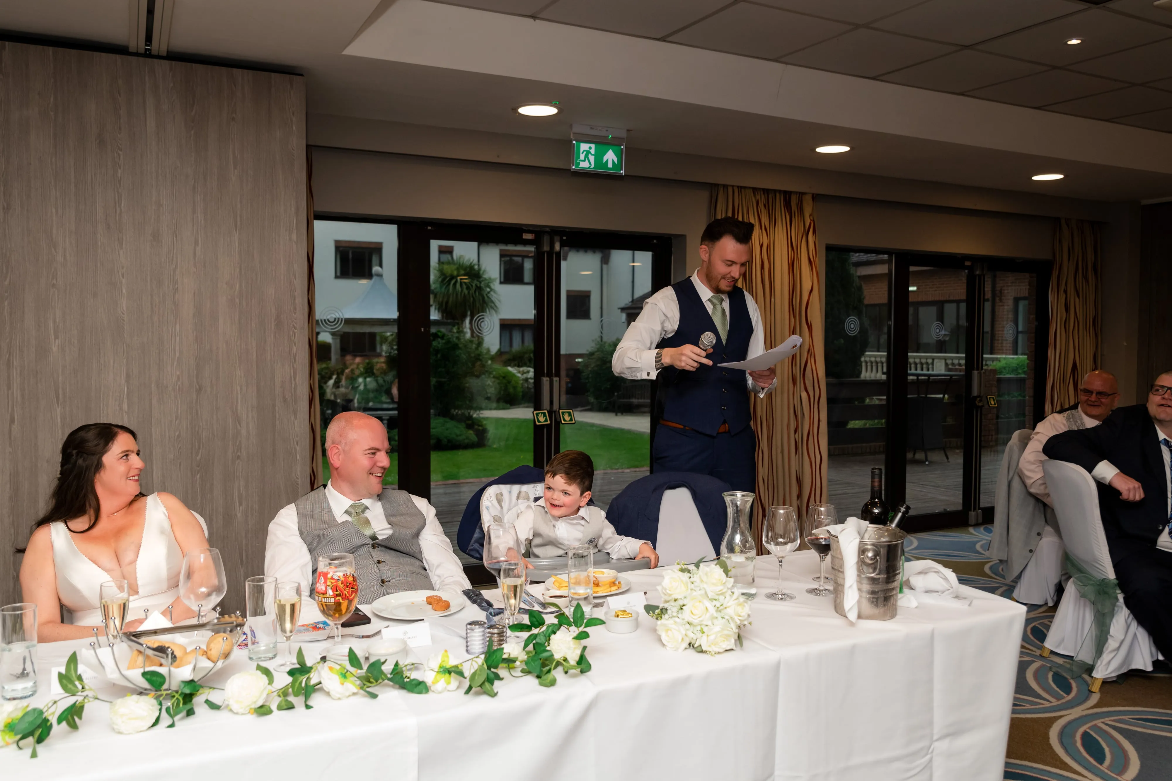 Best man stands at the top table delivering his speech with microphone, bride and groom seated and listening, full reception room visible behind