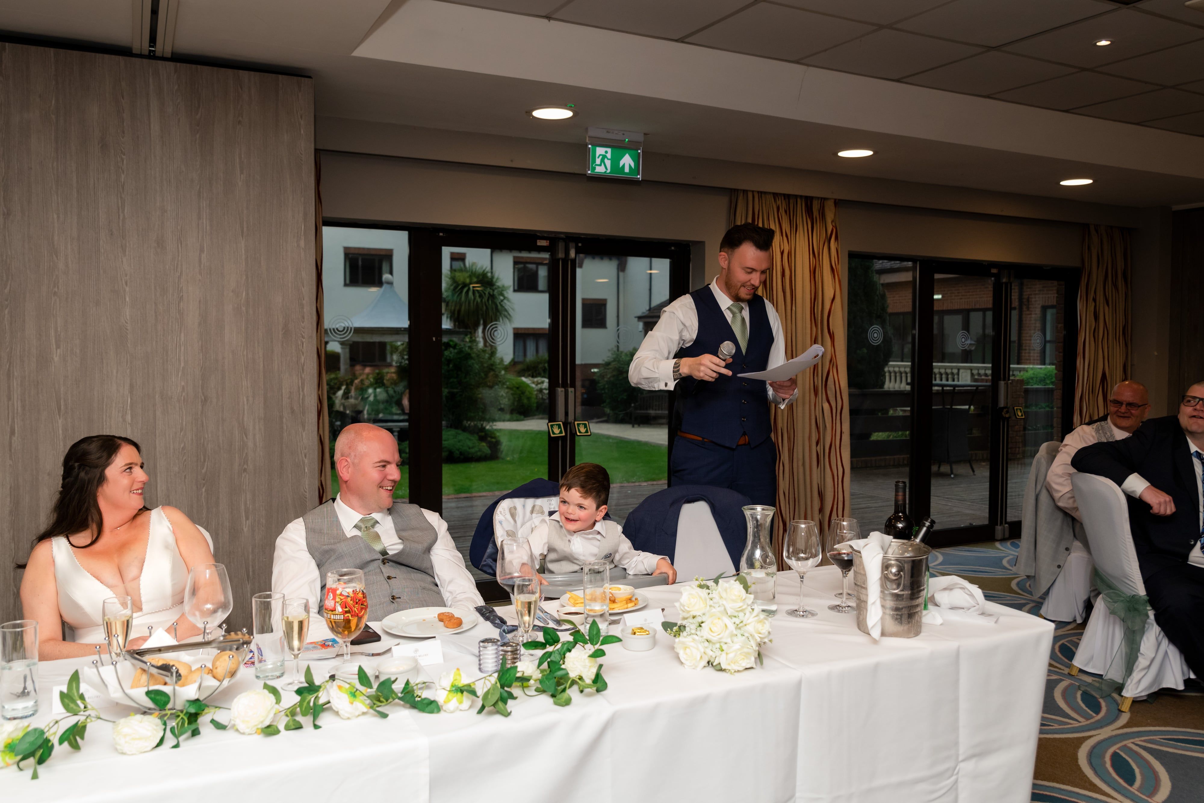 Best man stands at the top table delivering his speech with microphone, bride and groom seated and listening, full reception room visible behind