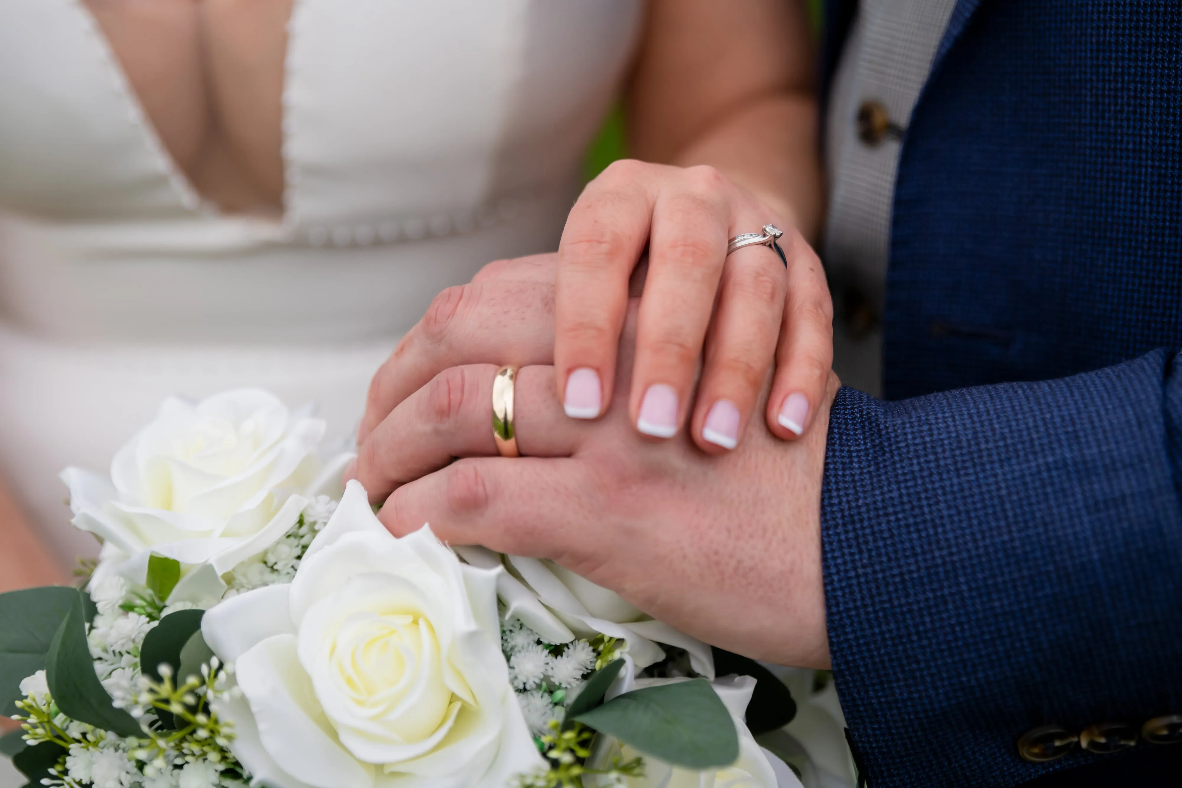 Close-up of bride and groom hands with wedding rings resting on white rose bouquet