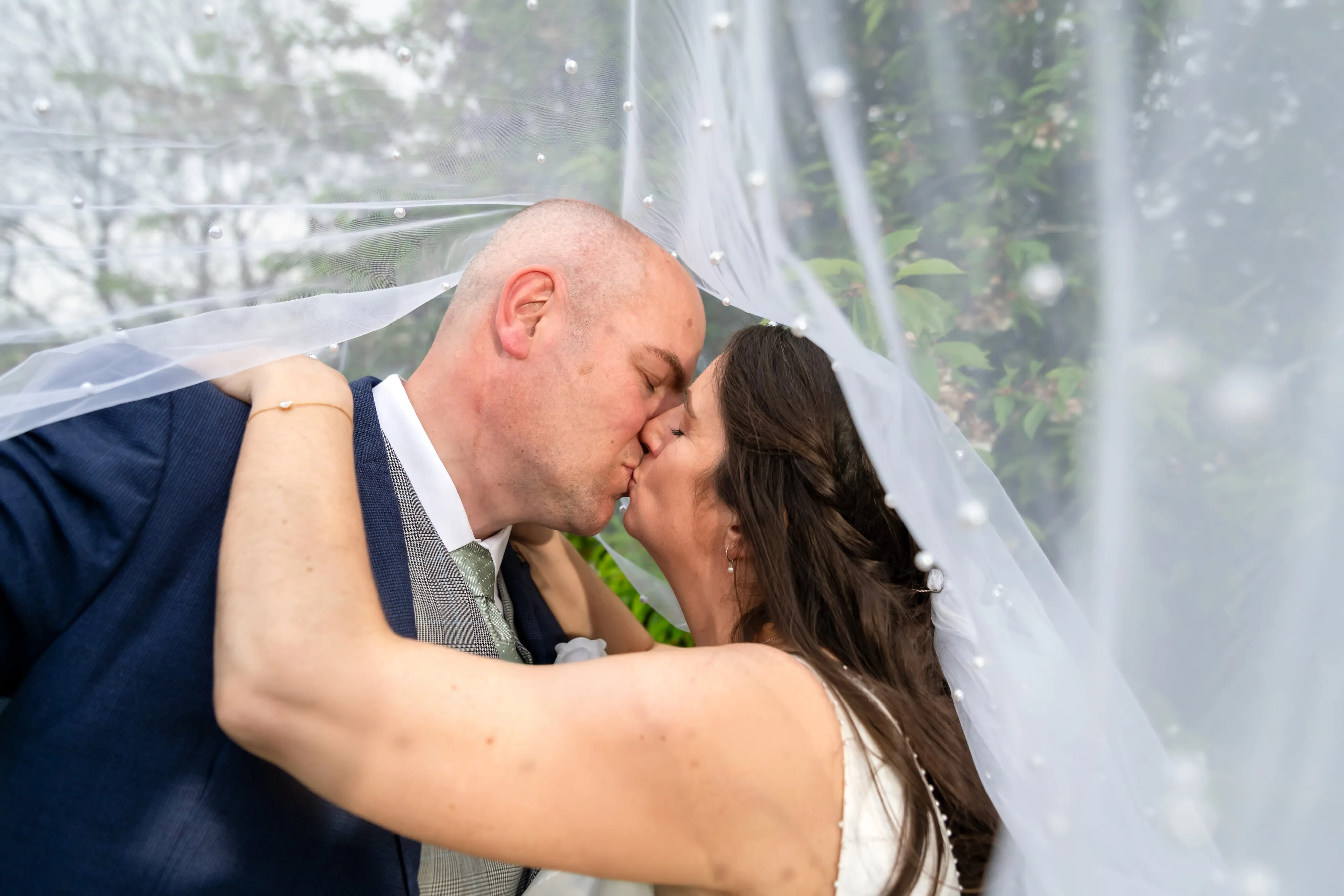 Close-up kiss of bride and groom beneath a pearl-studded sweeping veil, hedgerow softly out of focus behind