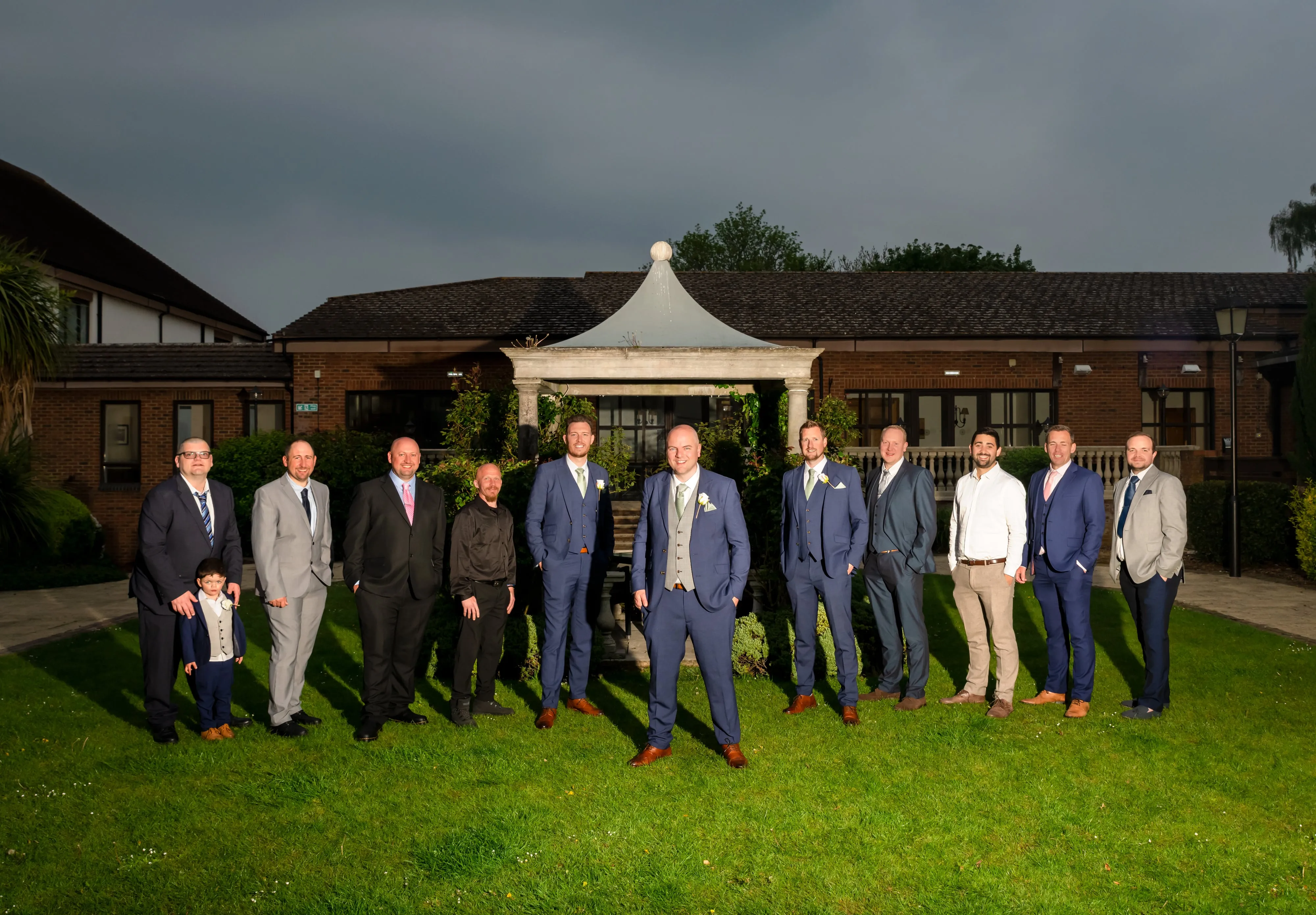 Groomsmen and family men line up in front of the stone pavilion at the Belfry under a moody twilight sky